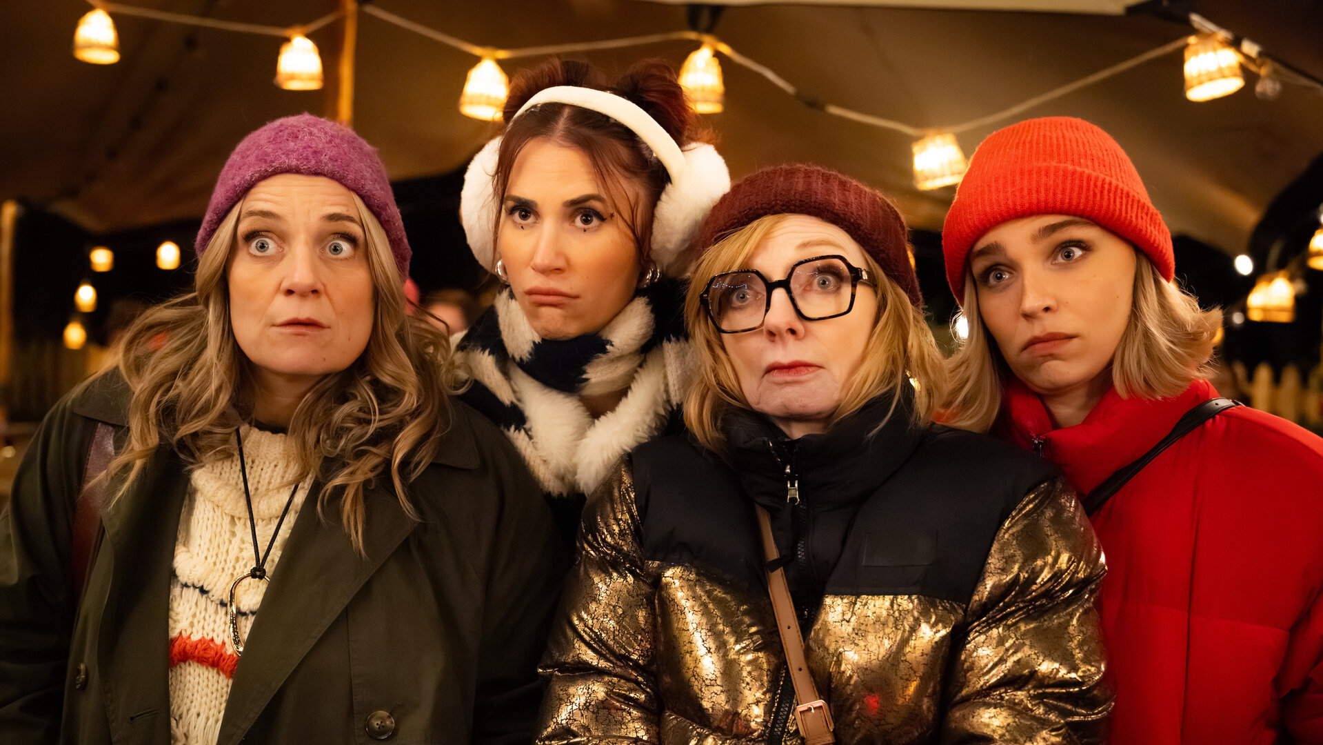 "Single Bells - Only Losers for Annie? - Herwig": Babs (Ruth Beeckmans), Daffy (Lize Feryn), Annie (Marleen Merckx) und Mina (Julie Van den Steen) sind mit Winterjacken und Mützen bekleidet und stehen in einem Zelt vor einem Holztisch mit  rot brennender Kerze. An der Decke hängt eine Lichterkette, im Hintergrund stehen Tische, auf denen rot leuchtende Kerzen stehen.