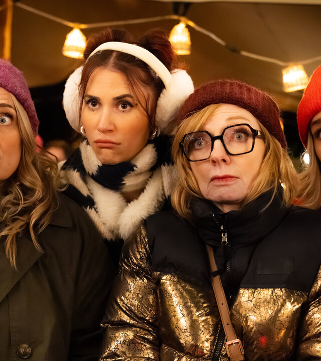 "Single Bells - Only Losers for Annie? - Herwig": Babs (Ruth Beeckmans), Daffy (Lize Feryn), Annie (Marleen Merckx) und Mina (Julie Van den Steen) sind mit Winterjacken und Mützen bekleidet und stehen in einem Zelt vor einem Holztisch mit  rot brennender Kerze. An der Decke hängt eine Lichterkette, im Hintergrund stehen Tische, auf denen rot leuchtende Kerzen stehen.