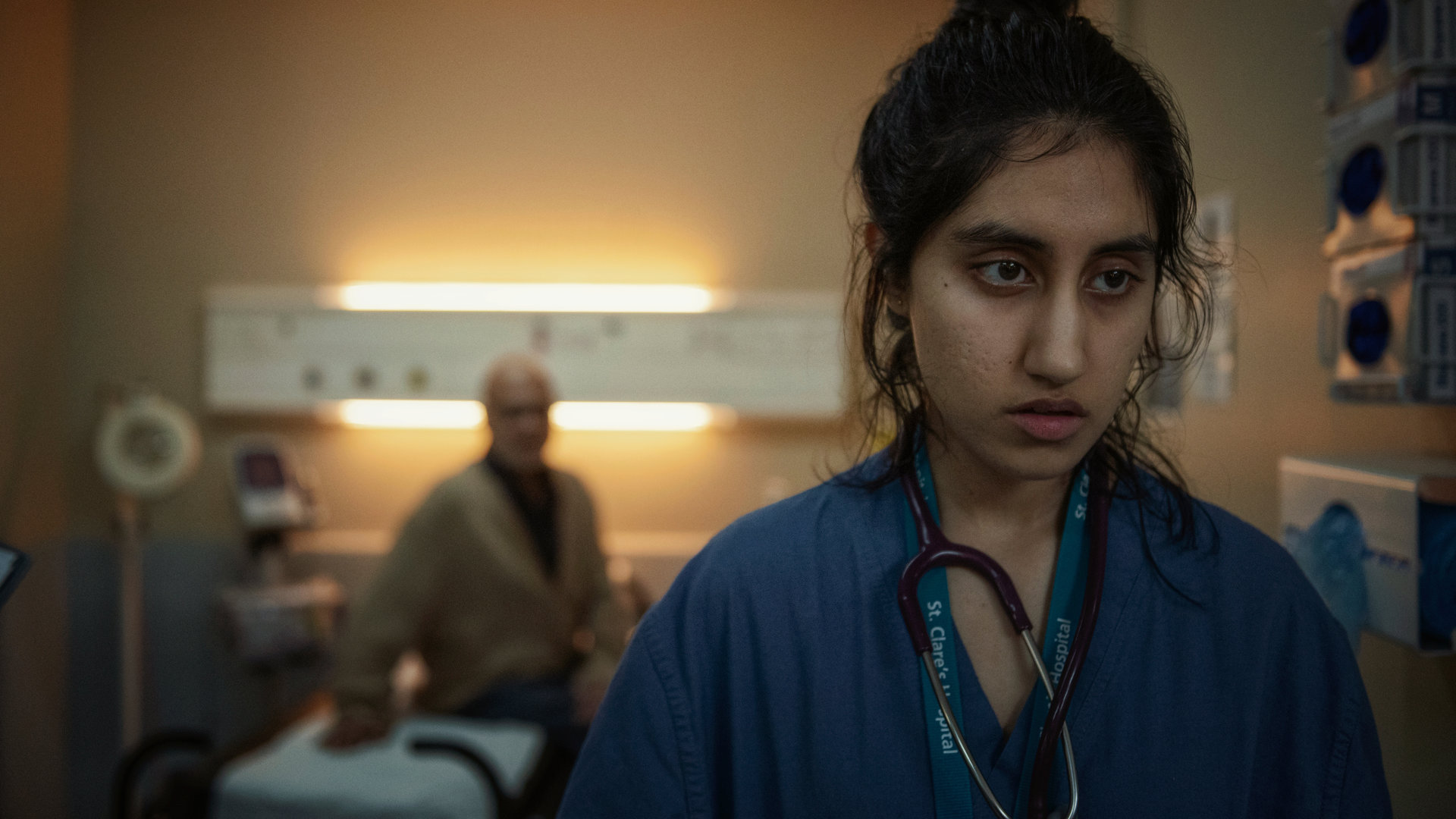 "This Is Going To Hurt - Shrutis Entscheidung": Shruti (Ambika Mod) steht in einem Krankenzimmer und schaut antriebslos in die Ferne, während ein Patient (Kalim Janjua) sie im Hintergrund beobachtet.