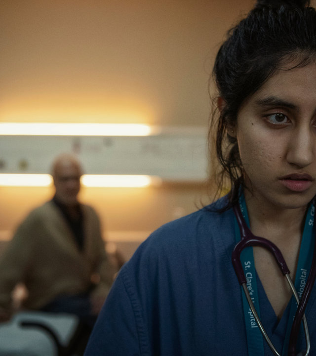 "This Is Going To Hurt - Shrutis Entscheidung": Shruti (Ambika Mod) steht in einem Krankenzimmer und schaut antriebslos in die Ferne, während ein Patient (Kalim Janjua) sie im Hintergrund beobachtet.