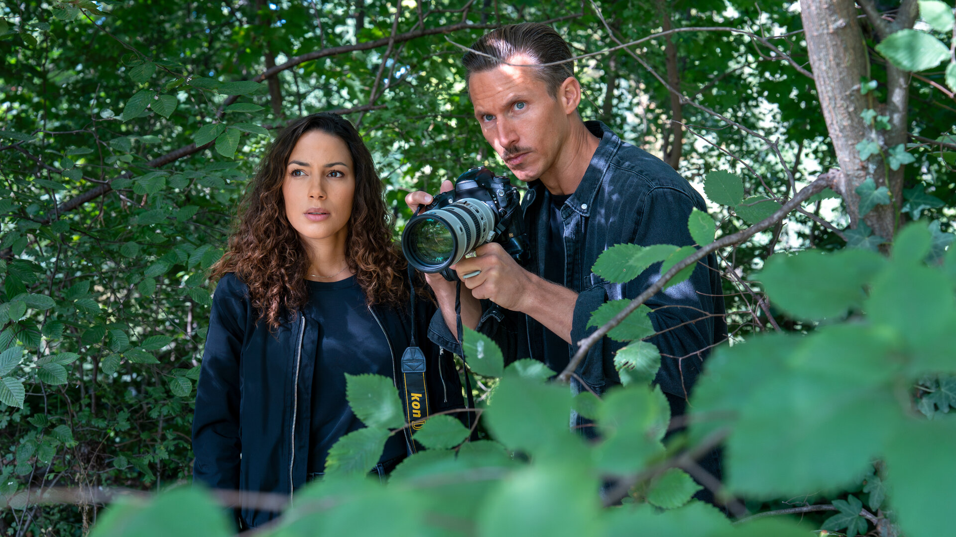 "SOKO Leipzig - Die Zeit heilt alle Wunden": Kim Nowak (Amy Mußul) und Moritz Brenner (Johannes Hendrik Langer) stehen nebeneinander in einem Gebüsch. Moritz hält eine Kamera und beide sehen ernst aus.