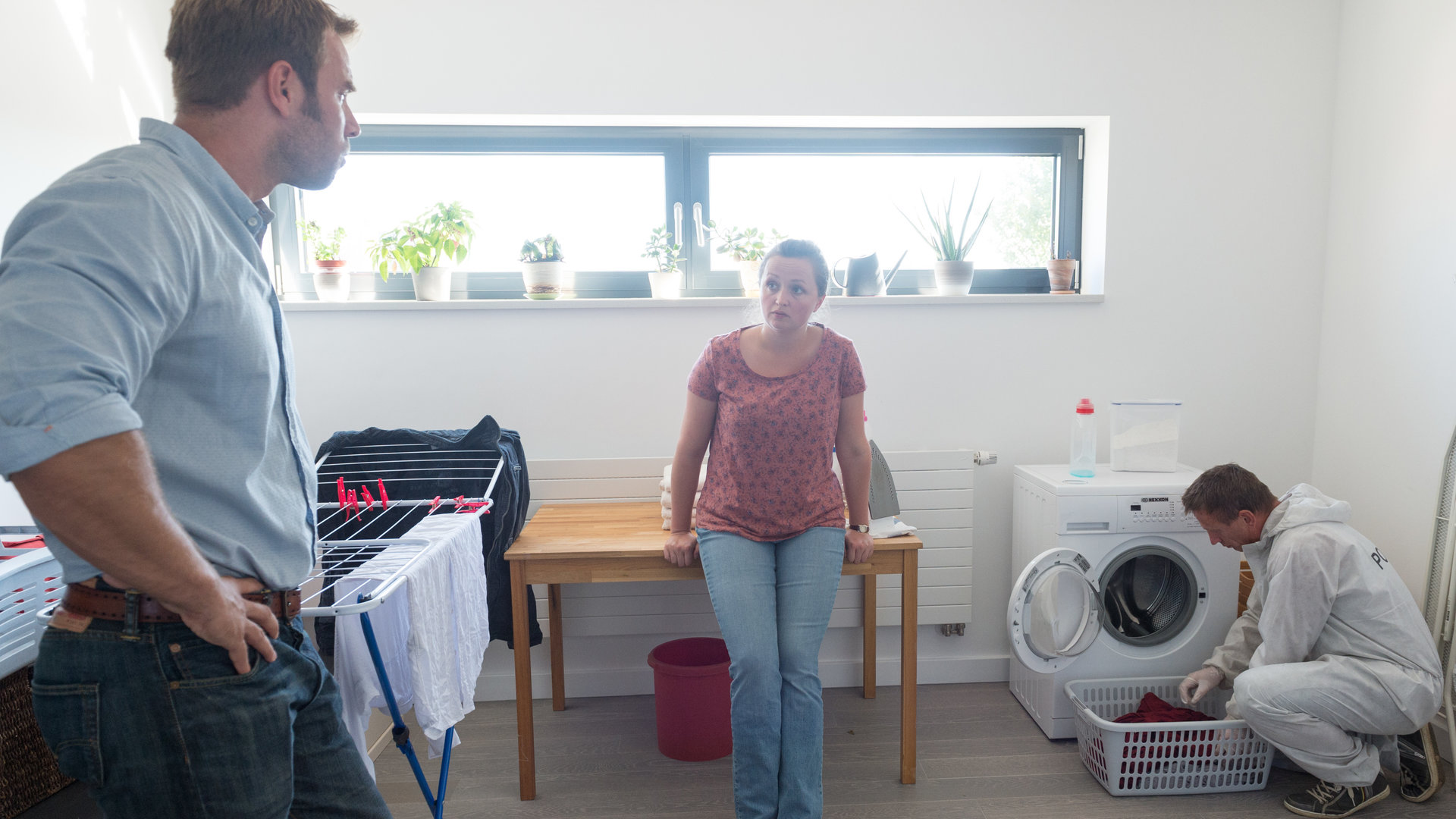 „SOKO Wismar – Klub der Aufreißer“: Lars Pöhlmann (Dominic Boeer) steht neben einem Wäscheständer und guckt zur Putzfrau Paula Sanders (Monika Oschek). Ein KTU-ler kniet neben der Waschmaschine und untersucht die Wäsche.