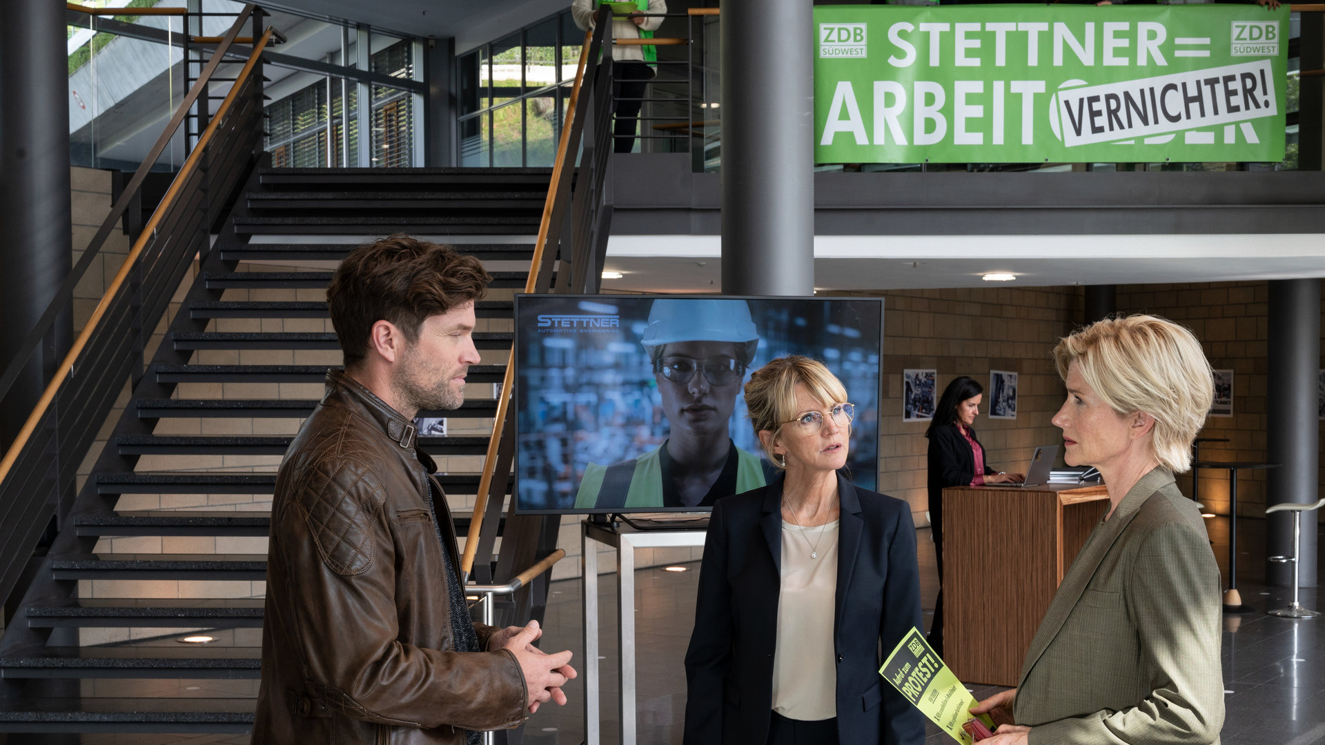 "SOKO Stuttgart – Unter Strom": Kriminalhauptkommissarin Martina Seiffert (Astrid M. Fünderich) und Kriminalhauptkommissar Jo Stoll (Peter Ketnath) befragen Anette Stettner (Esther Esche) im Foyer Stuttgarter Traditionsunternehmen Stettner Werke. Im Hintergrund sieht man eine große Treppe und eine Empfangsdame, die an einem Empfangstresen steht. Martina hält einen Flyer in den Händen, auf dem man das Wort "Protest" lesen kann. An dem Geländer der Treppe hängt ein Protestplakat.