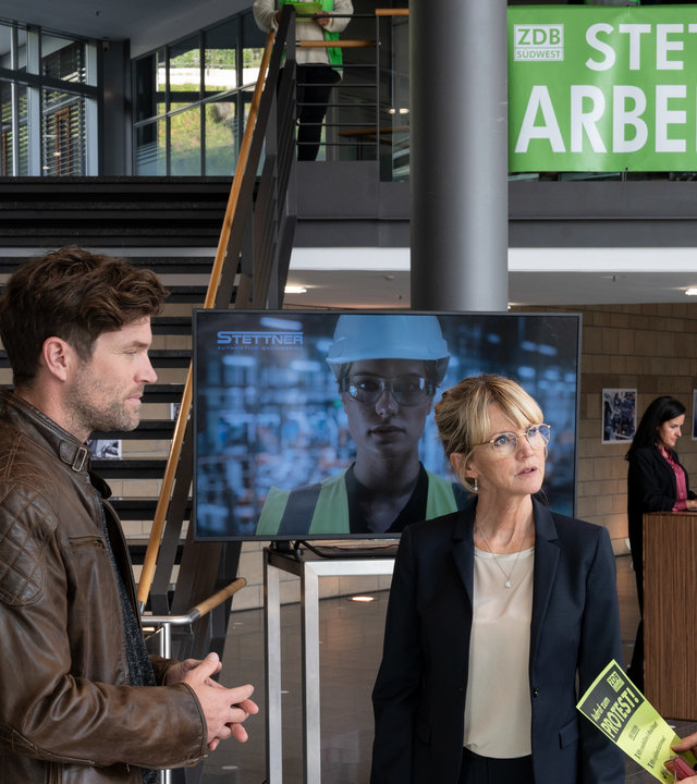 "SOKO Stuttgart – Unter Strom": Kriminalhauptkommissarin Martina Seiffert (Astrid M. Fünderich) und Kriminalhauptkommissar Jo Stoll (Peter Ketnath) befragen Anette Stettner (Esther Esche) im Foyer Stuttgarter Traditionsunternehmen Stettner Werke. Im Hintergrund sieht man eine große Treppe und eine Empfangsdame, die an einem Empfangstresen steht. Martina hält einen Flyer in den Händen, auf dem man das Wort "Protest" lesen kann. An dem Geländer der Treppe hängt ein Protestplakat.