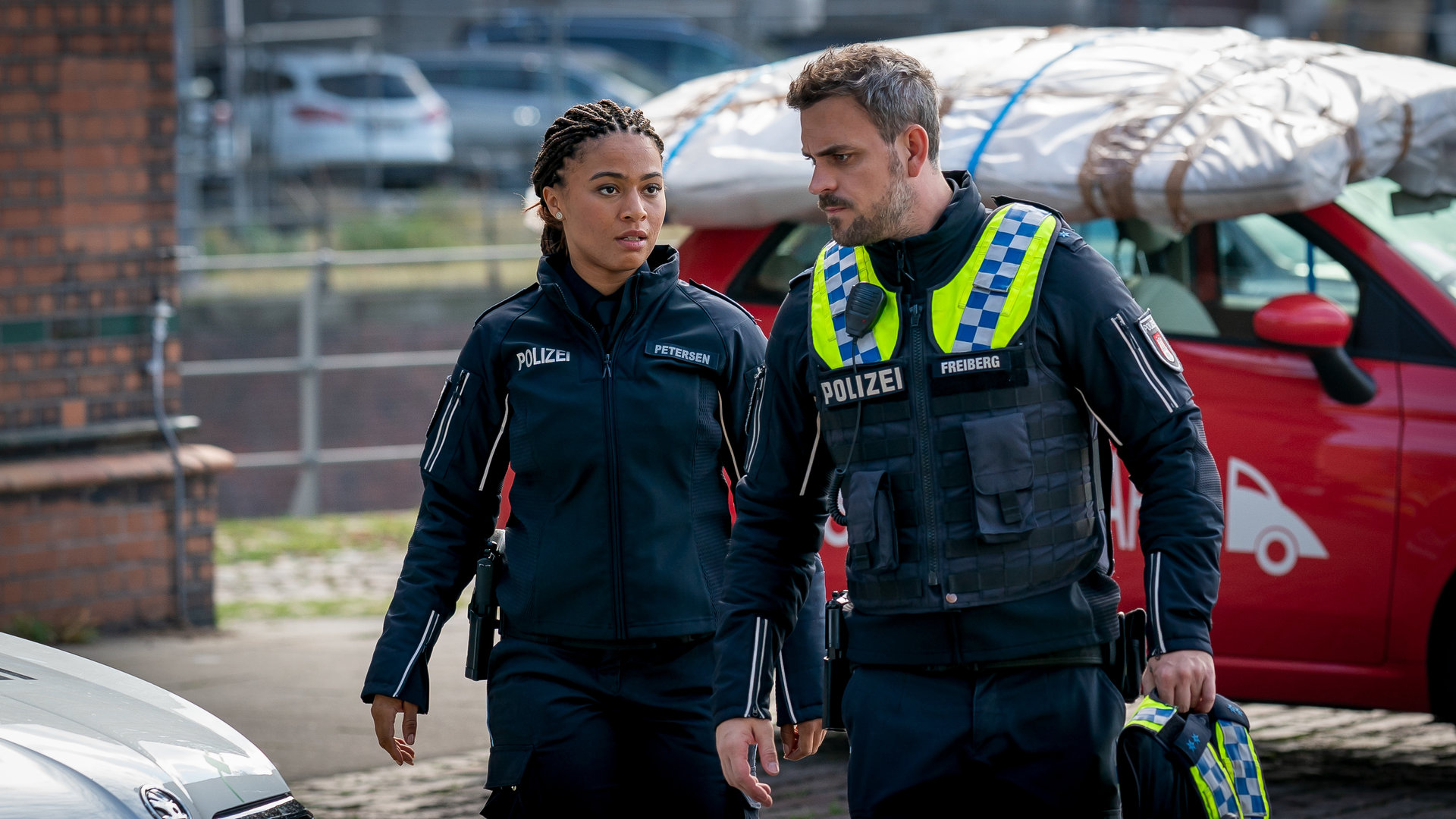 "Notruf Hafenkante - Ausgemustert": Die Polizistin Daisy Petersen (Aysha Joy Samuel) und ihr Kollege Kris Freiberg (Marc Barthel) gehen nebeneinander an parkenden Autos vorbei.