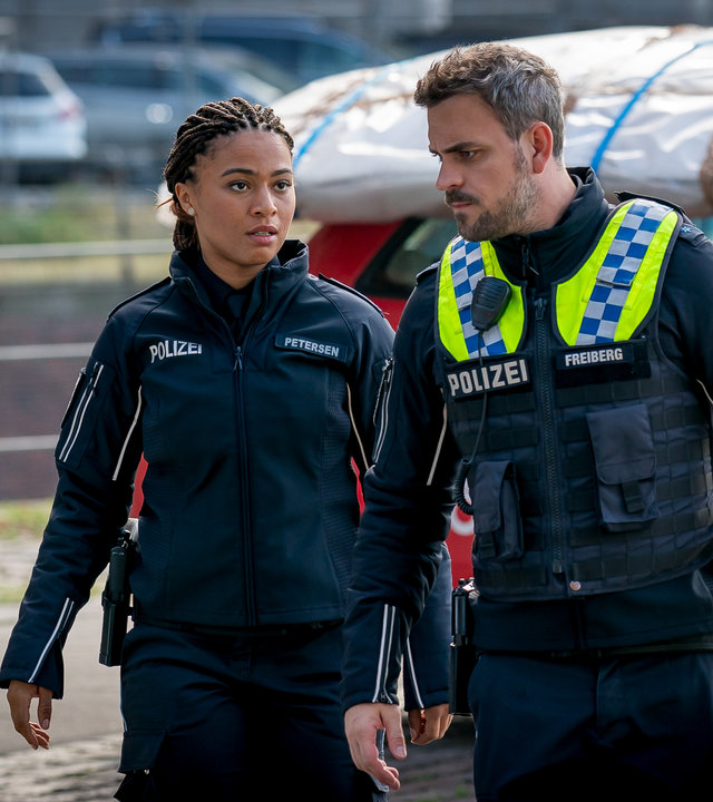 "Notruf Hafenkante - Ausgemustert": Die Polizistin Daisy Petersen (Aysha Joy Samuel) und ihr Kollege Kris Freiberg (Marc Barthel) gehen nebeneinander an parkenden Autos vorbei.