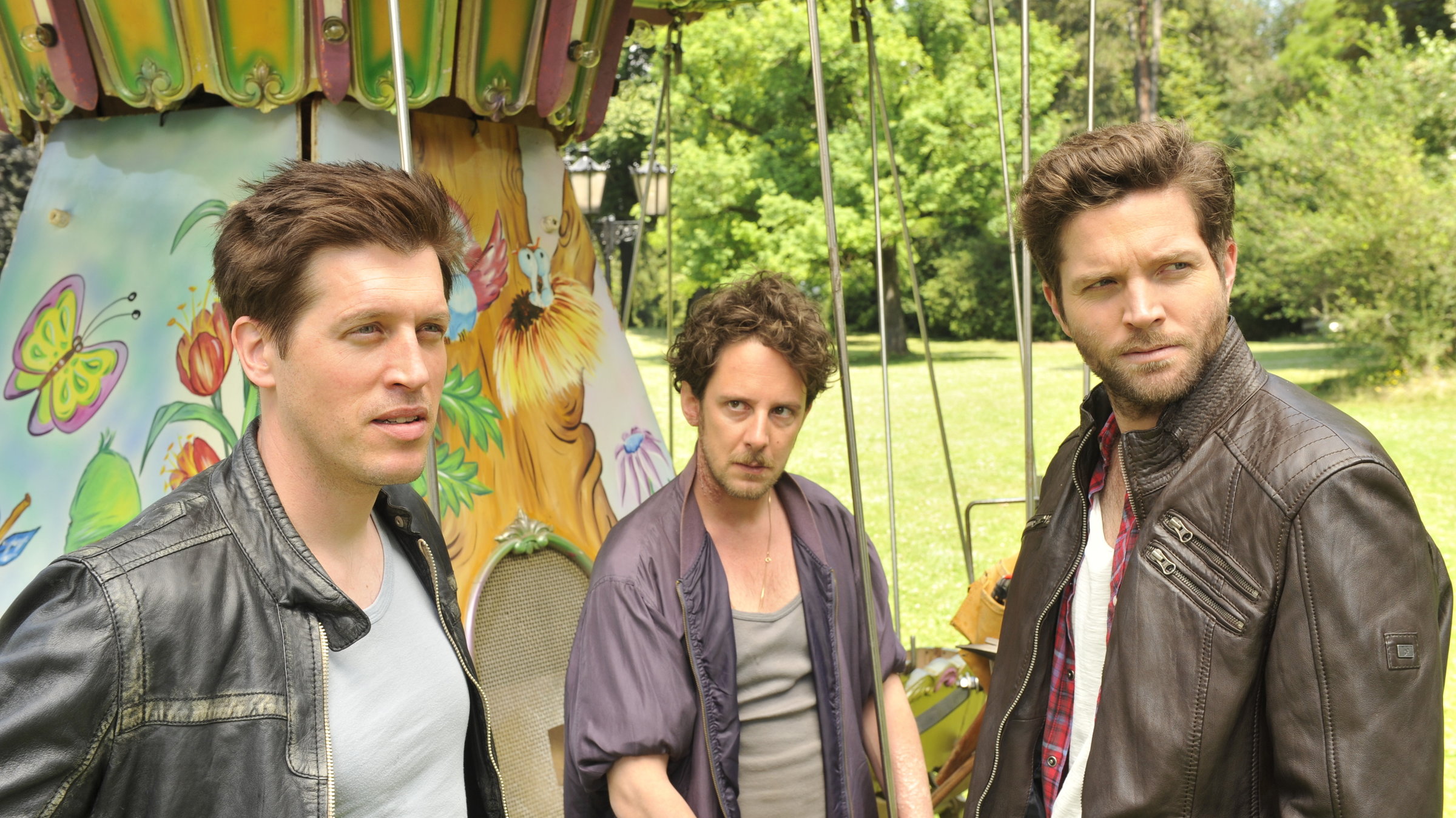 "SOKO Stuttgart - Rad der Zeit": Jo Stoll (Peter Ketnath) steht mit den Brüdern Levi Horwitz (Nic Romm) und Kaspar Horwitz (Marian Meder) auf einer grünen Wiese im Park vor einem alten Kinderkarussell.