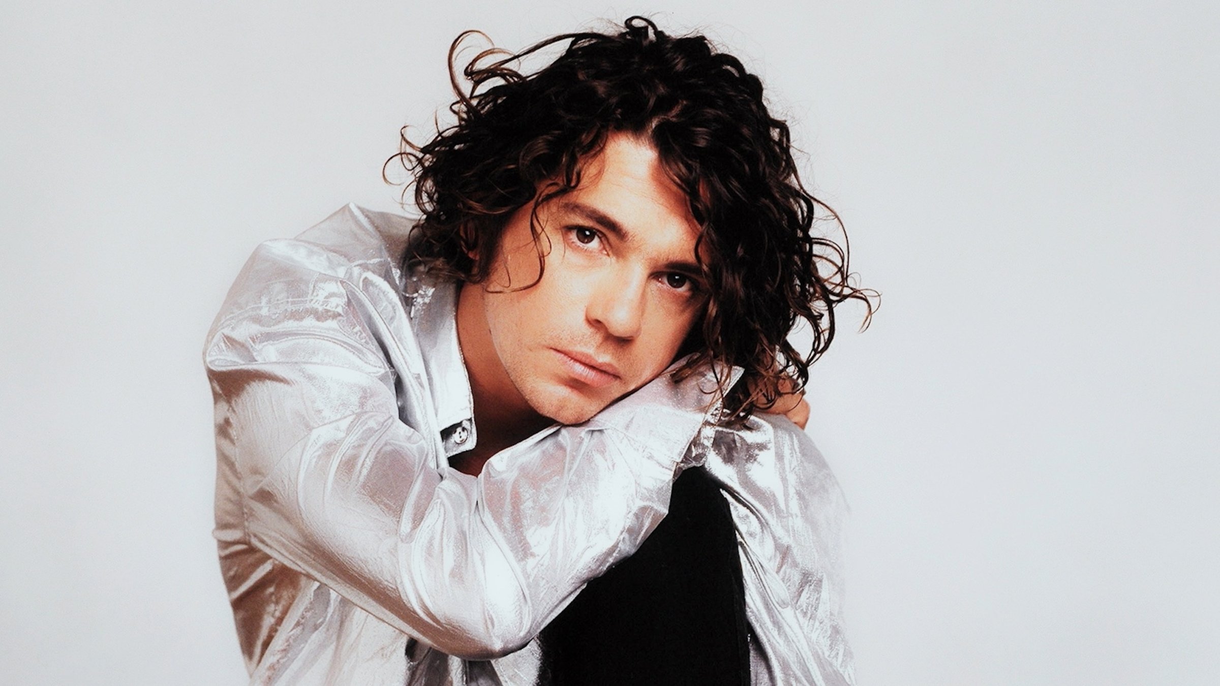 "The Day the Rock Star Died: Michael Hutchence": Frontalaufnahme vom knieenden Michael Hutchence. Er trägt ein schimmerndes weißes Hemd und stützt den Kopf auf dem Knie ab.