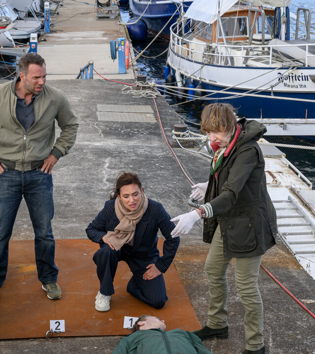 "SOKO Wismar – Im Wahn der Ahnen": Am Tatort am Flussufer haben sich die Kriminalhauptkommissare Lars Pöhlmann (Dominic Boeer) und Karoline Joost (Nike Fuhrmann) mit Rechtsmedizinerin Dr. Helene Sturbeck (Katharina Blaschke) um die Leiche des Informatikstudenten Konrad Kessler (Kleindarsteller) versammelt. Neben dem Tatort liegen einige Schiffe vertäut.