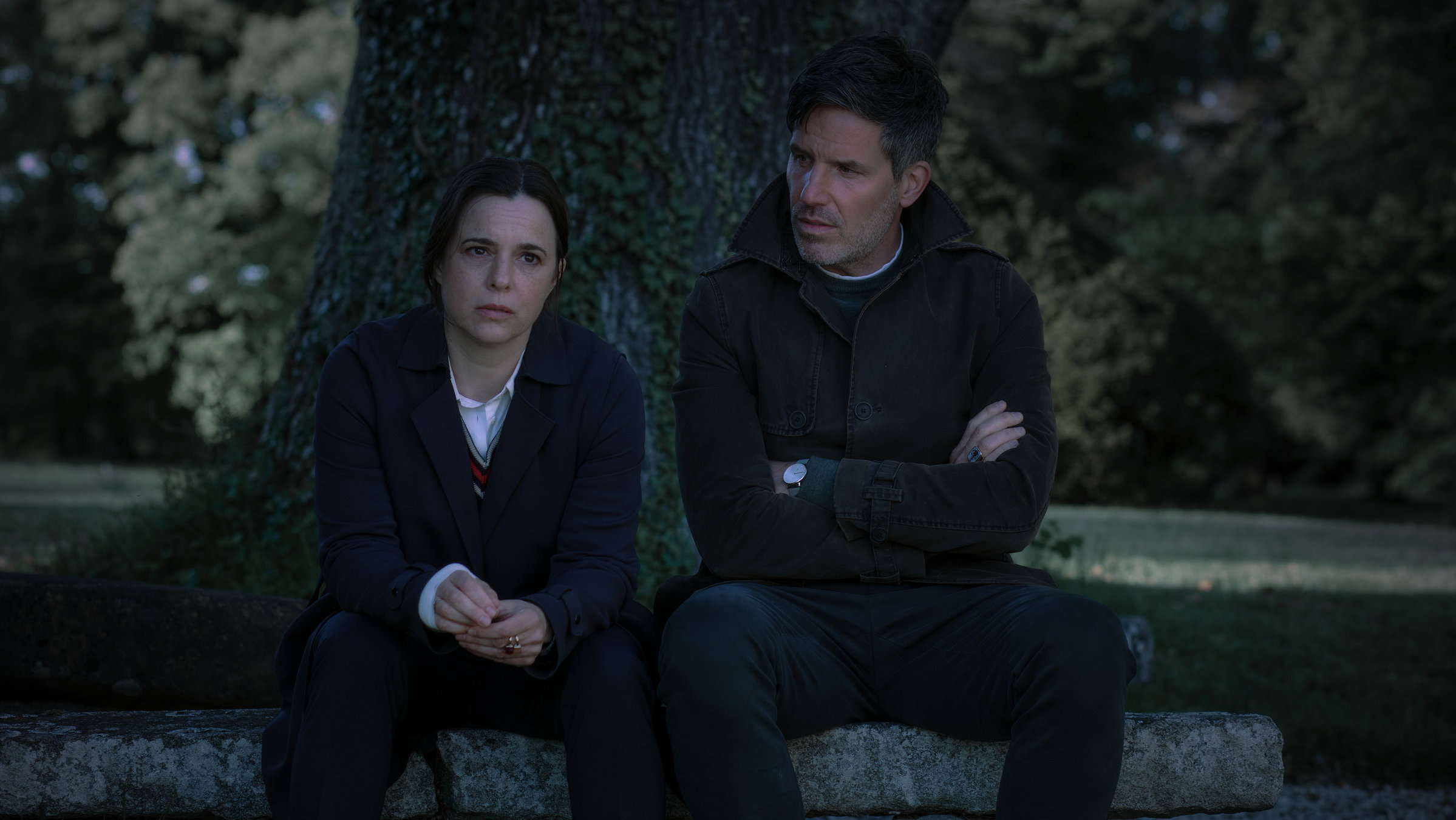"Muttertag (2) - Ein Taunuskrimi": Pia Sander (Annika Kuhl) und Oliver von Bodenstein (Tim Bergmann) sitzen im Halbdunkel auf einem großen Stein unter einem großen Baumstamm. Pia stützt ihre Arme auf den Knien ab und blickt angespannt in die Ferne, Oliver schaut zu seiner Kollegin. Er hat die Arme verschränkt.