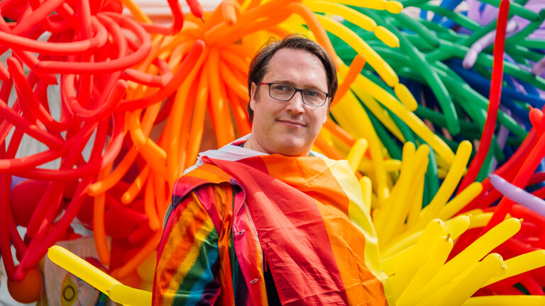 "einfach Mensch": Martin Bühren: Queer mit Behinderung" Martin Bühren auf einer Wiese mit einer Regenbogenflagge