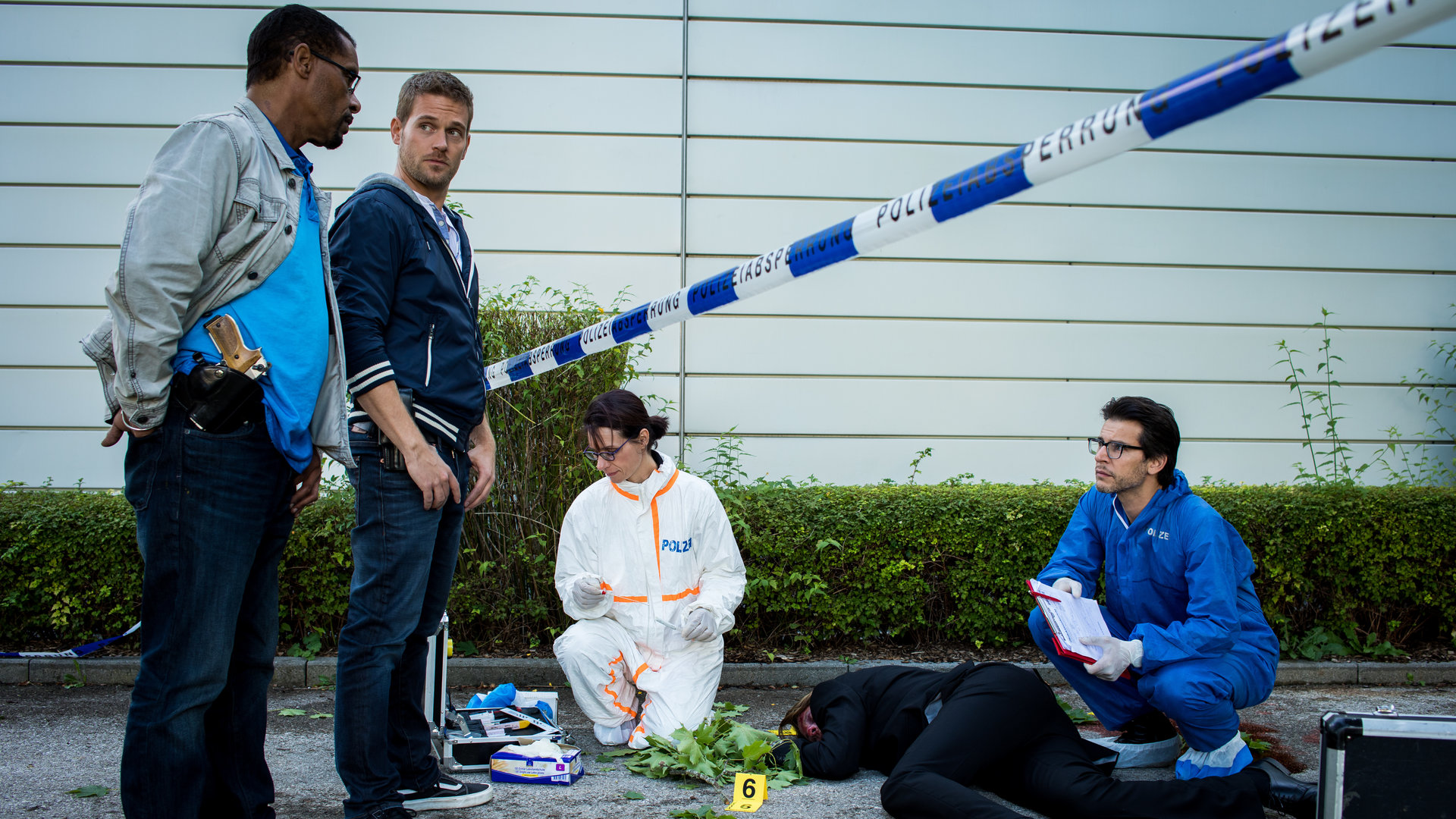 "SOKO München - Es braut sich was zam": Franz Ainfachnur (Christofer v. Beau) und Dominik Morgenstern (Joscha Kiefer) stehen nebeneinander vor einem polizeilichen Absperrband am Tatort. Dort arbeiten eine Kollegin der Spurensicherung und Dr. Weissenböck (Florian Odendahl), der in der  Hocke neben der am Boden liegenden Leiche sitzt.