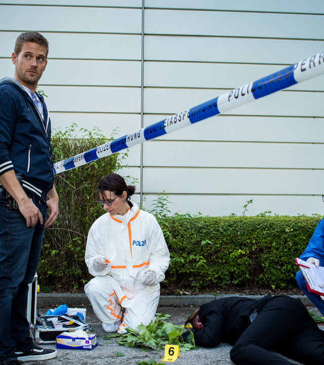 "SOKO München - Es braut sich was zam": Franz Ainfachnur (Christofer v. Beau) und Dominik Morgenstern (Joscha Kiefer) stehen nebeneinander vor einem polizeilichen Absperrband am Tatort. Dort arbeiten eine Kollegin der Spurensicherung und Dr. Weissenböck (Florian Odendahl), der in der  Hocke neben der am Boden liegenden Leiche sitzt.