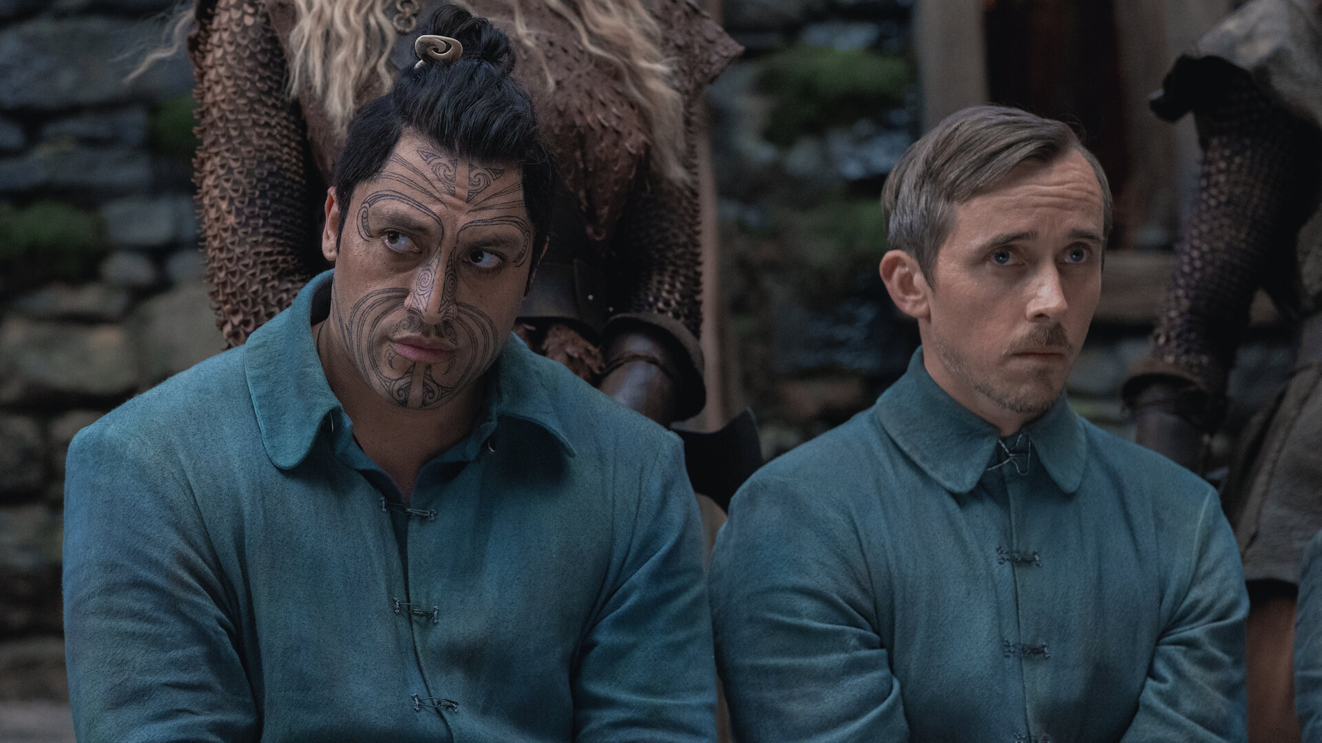 "Nautilus - Ein Schatz": Kai (Tyrone Ngatai) und Cuff (Benedict Hardie) sitzen nebeneinander im Freien. Sie tragen einheitliche Kleidung. Hinter ihnen stehen im Anschitt zwei Frauen (Komparsinnen).