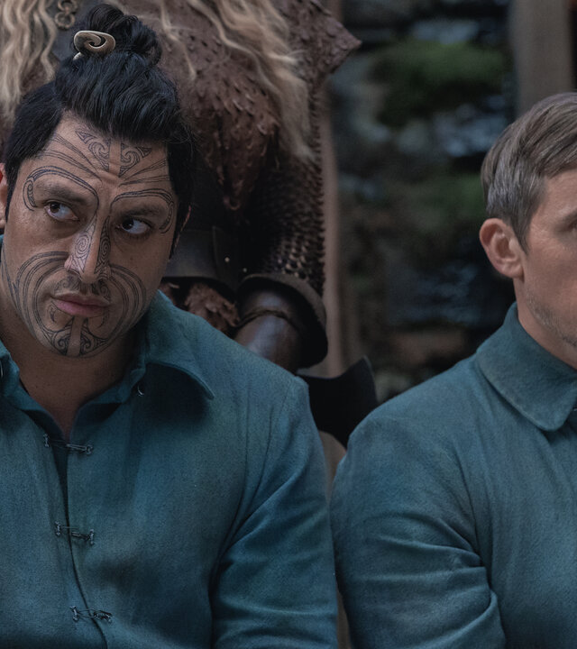 "Nautilus - Ein Schatz": Kai (Tyrone Ngatai) und Cuff (Benedict Hardie) sitzen nebeneinander im Freien. Sie tragen einheitliche Kleidung. Hinter ihnen stehen im Anschitt zwei Frauen (Komparsinnen).