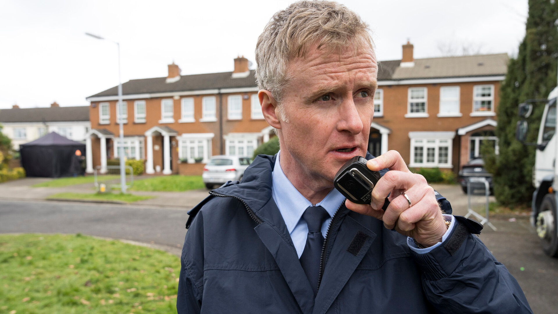 "Harry Wild - Mörderjagd in Dublin - Der Feind in meiner Soap": Frank Stynes (Damian Kearney) steht auf der Straße und spricht in ein Funkgerät. Im Hintergrund stehen Reihenhäuser hinter einer Rasenfläche.