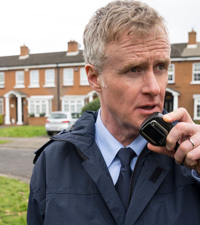 "Harry Wild - Mörderjagd in Dublin - Der Feind in meiner Soap": Frank Stynes (Damian Kearney) steht auf der Straße und spricht in ein Funkgerät. Im Hintergrund stehen Reihenhäuser hinter einer Rasenfläche.