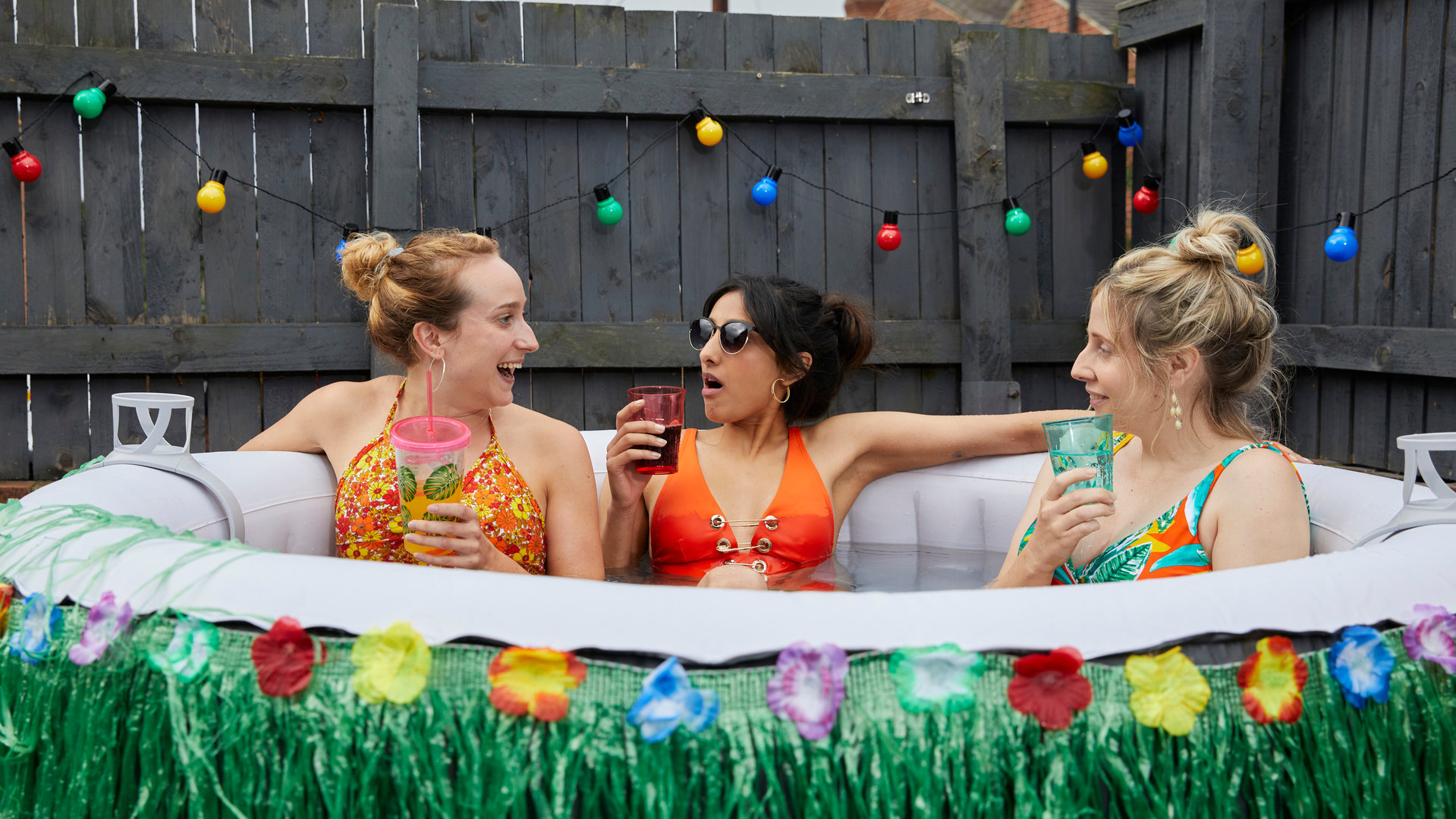 "Hullraisers - Breadcakes": Toni (Leah Brotherhead), Rana (Taj Atwal) und Paula (Sinead Matthews)  sitzen zusammen im Bikini in einem aufblasbaren Swimmingpool und schauen sich lachend an, während sie etwas trinken.