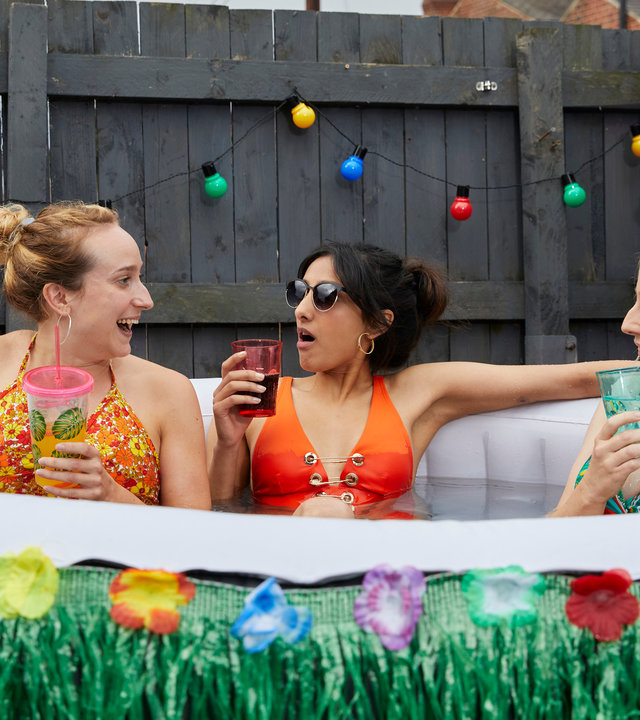 "Hullraisers - Breadcakes": Toni (Leah Brotherhead), Rana (Taj Atwal) und Paula (Sinead Matthews)  sitzen zusammen im Bikini in einem aufblasbaren Swimmingpool und schauen sich lachend an, während sie etwas trinken.