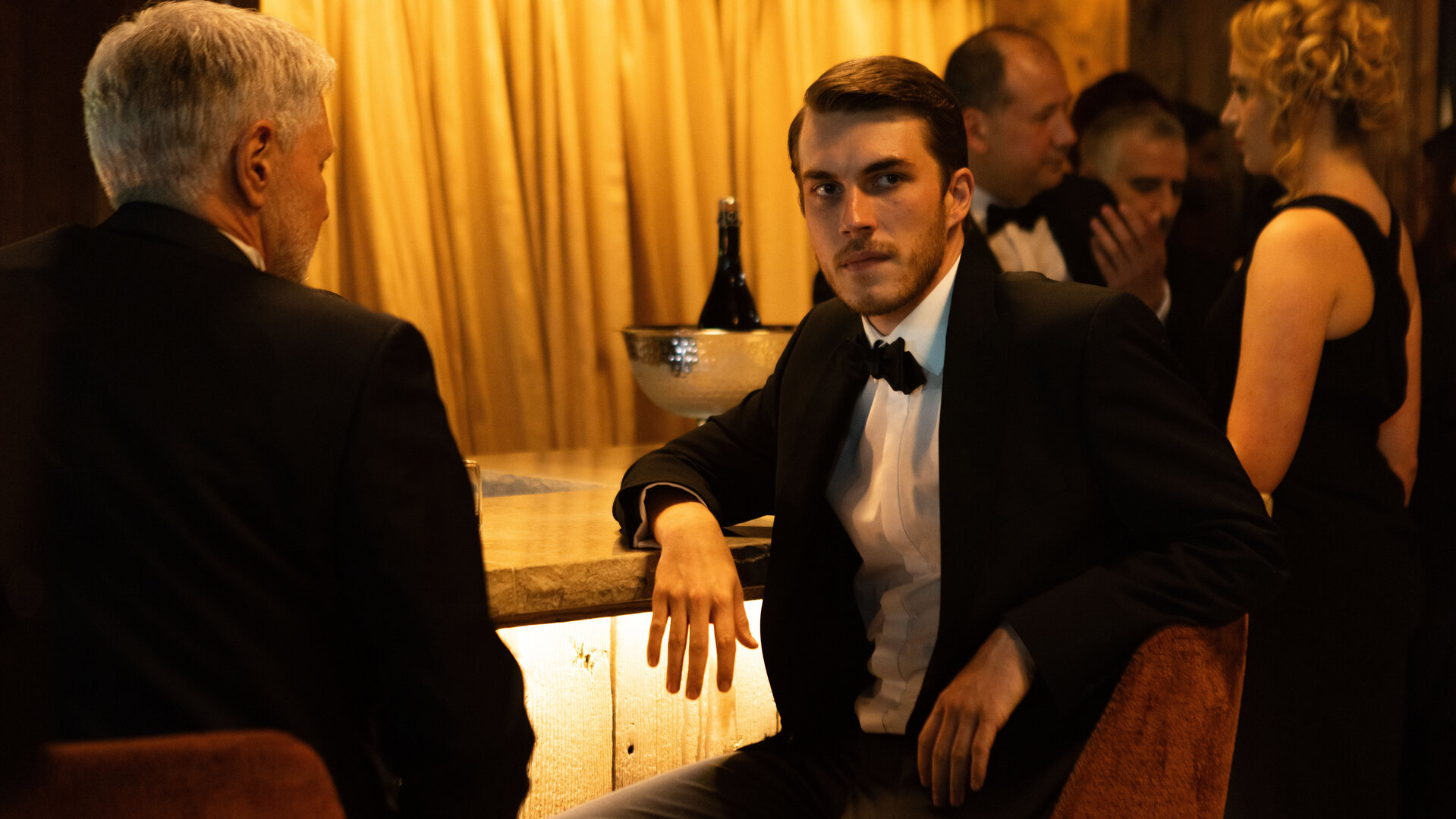 "The Kollective - Unkonventionelle Methoden": Aaron (Felix Mayr) sitzt an einer Bar. Er trägt einen dunklen Anzug mit Fliege. Um ihn herum befinden sich weitere elegant gekleidete Menschen (Komparsen).