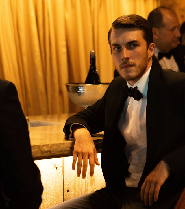 "The Kollective - Unkonventionelle Methoden": Aaron (Felix Mayr) sitzt an einer Bar. Er trägt einen dunklen Anzug mit Fliege. Um ihn herum befinden sich weitere elegant gekleidete Menschen (Komparsen).