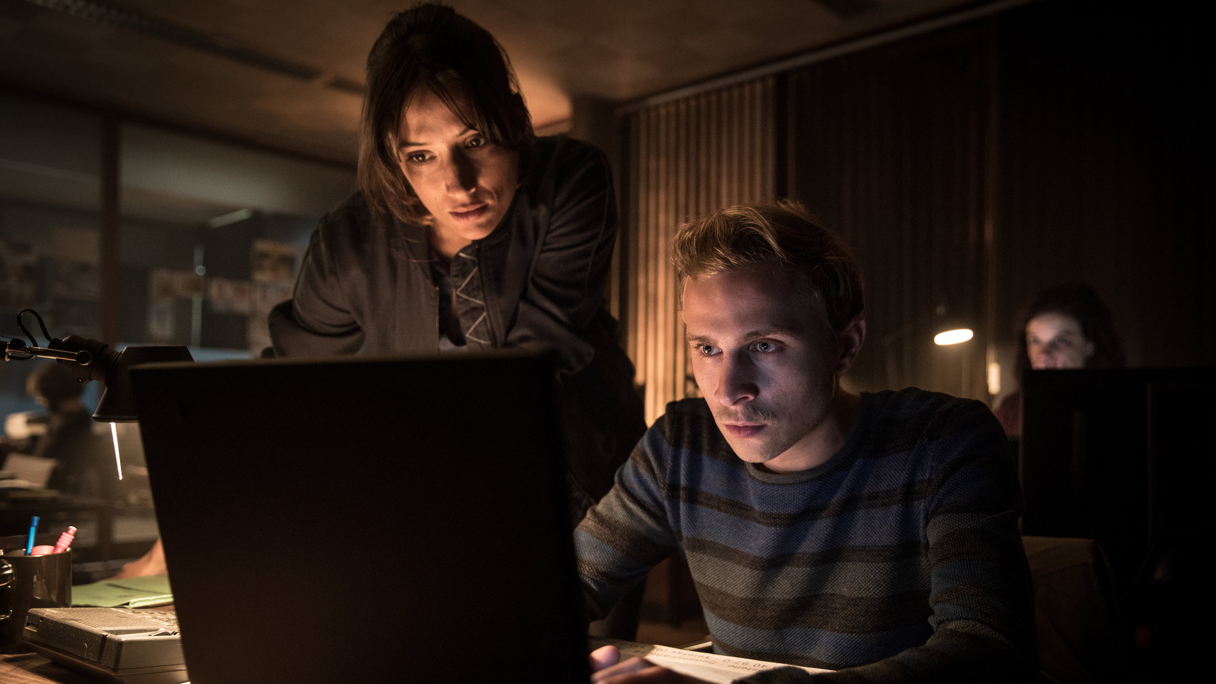 "The Team II (3)": Sabine (Melanie Winiger) steht neben einem Schreibtisch im abgedunkelten Polizeirevier, an dem Bastian (Max Hubacher) sitzt. Beide schauen auf den Bildschirm des Laptops, das vor Bastian auf dem Tisch steht.