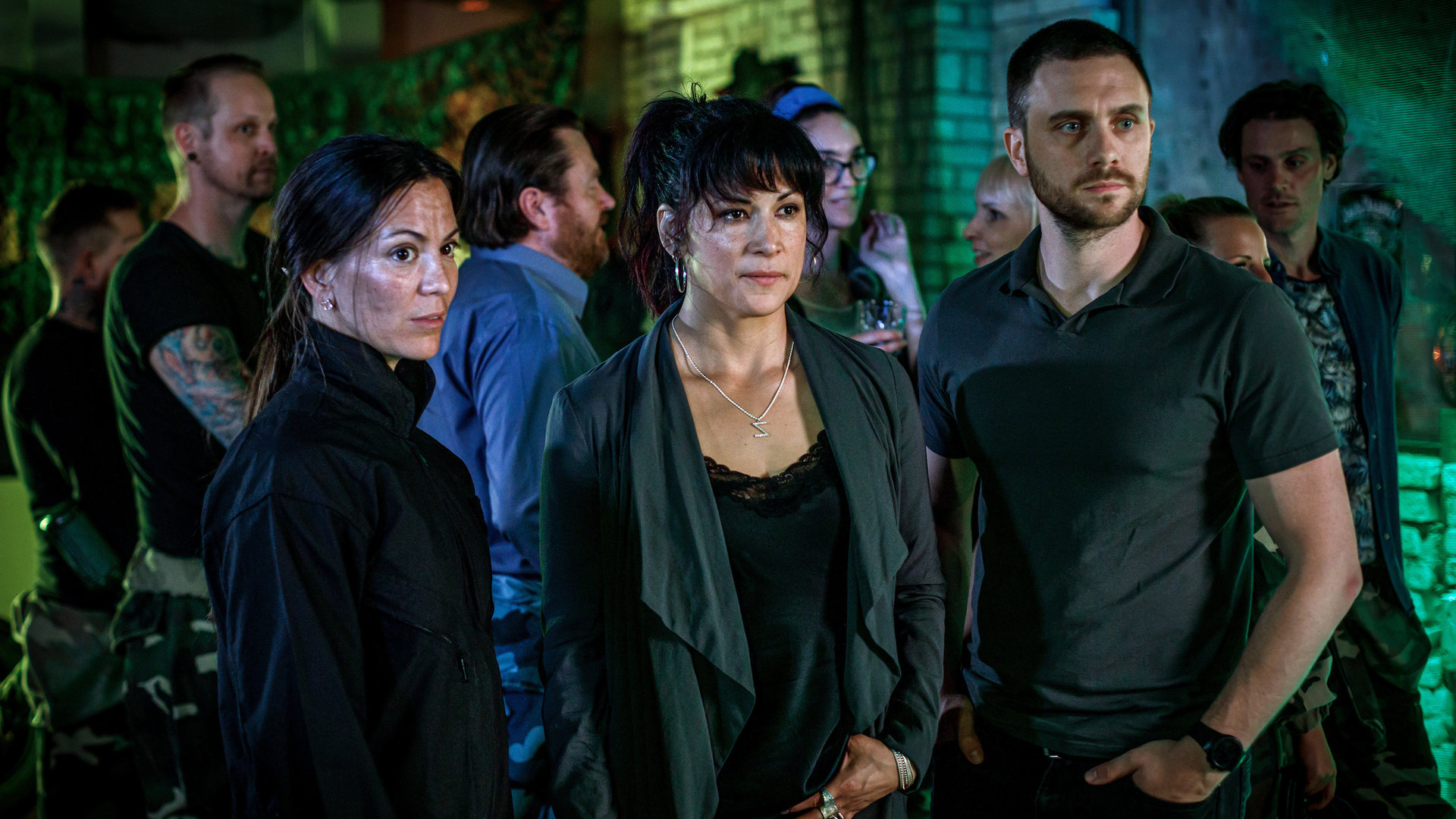 "Kommissar Beck: Rage Room": Åsa Karlsson (Johanna Lazcano), Cissi Karlsson (Malin Arvidsson) und Josef Eriksson (Martin Wallström) stehen in einer Halle für Adventure Games und blicken in eine Richtung. Hinter ihnen stehen weitere Gäste.