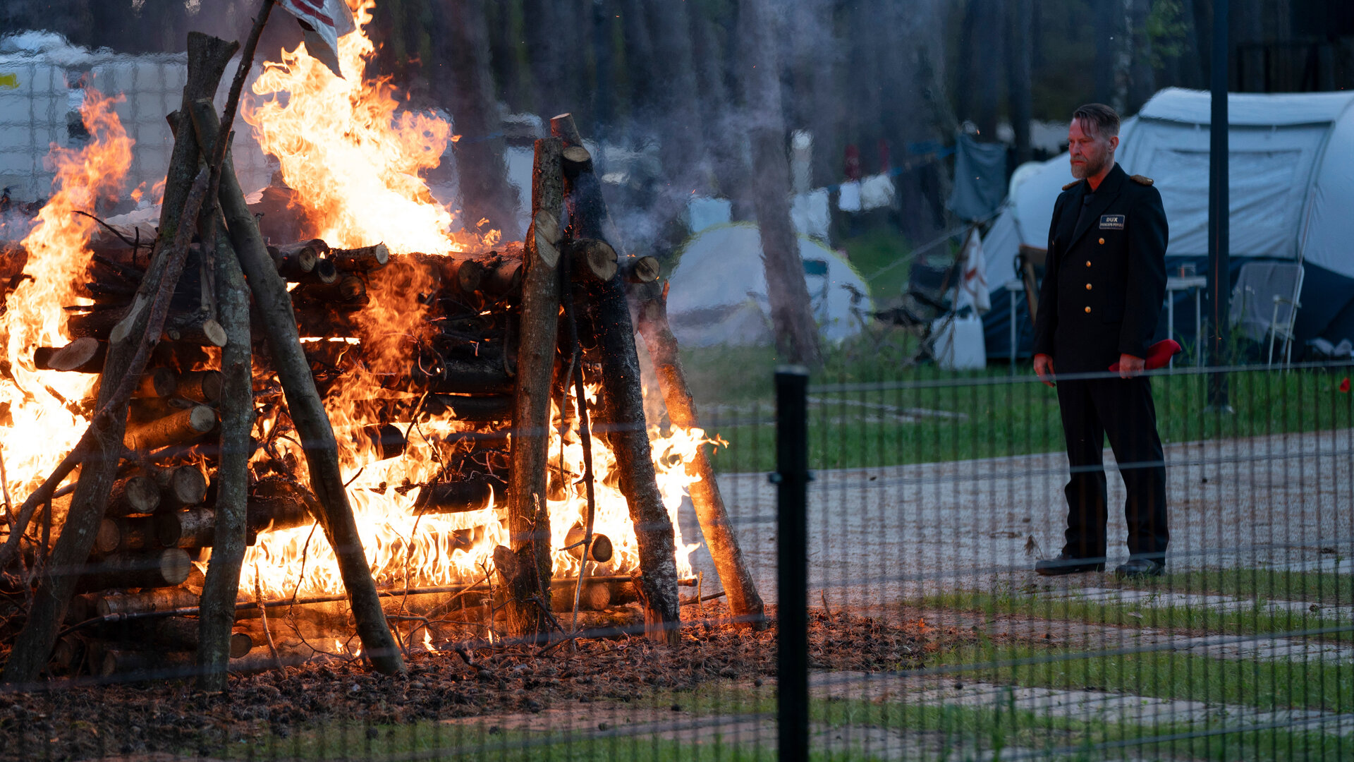 "Sløborn III – Die leise Nacht": Erik (Manuel Harder) steht in Uniform vor einem brennenden Holzunterschlupf und starrt in das Feuer. Im Hintergrund stehen Zelte auf einer Waldlichtung.
