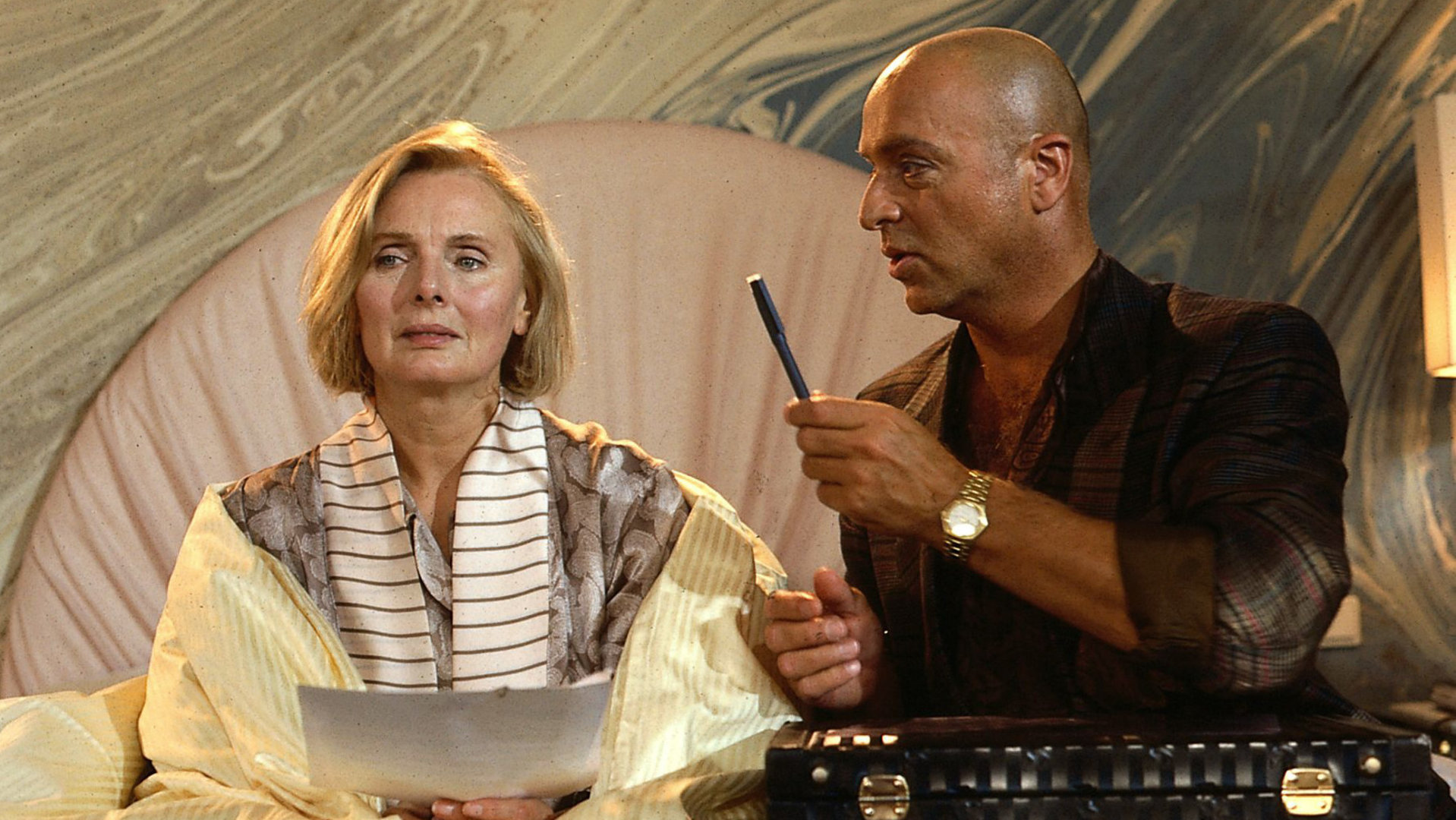 "Das Erbe der Guldenburgs - Der letzte Strohhalm": Achim (Wilfried Baasner) fordert von Margot (Ruth Maria Kubitschek) eine Unterschrift ein. Beide sitzen auf einem Bett, Achim hat einen schwarzen Aktenkoffer auf den Beinen liegen. Margot hält Formulare in der Hand, Achim reicht ihr einen Stift.