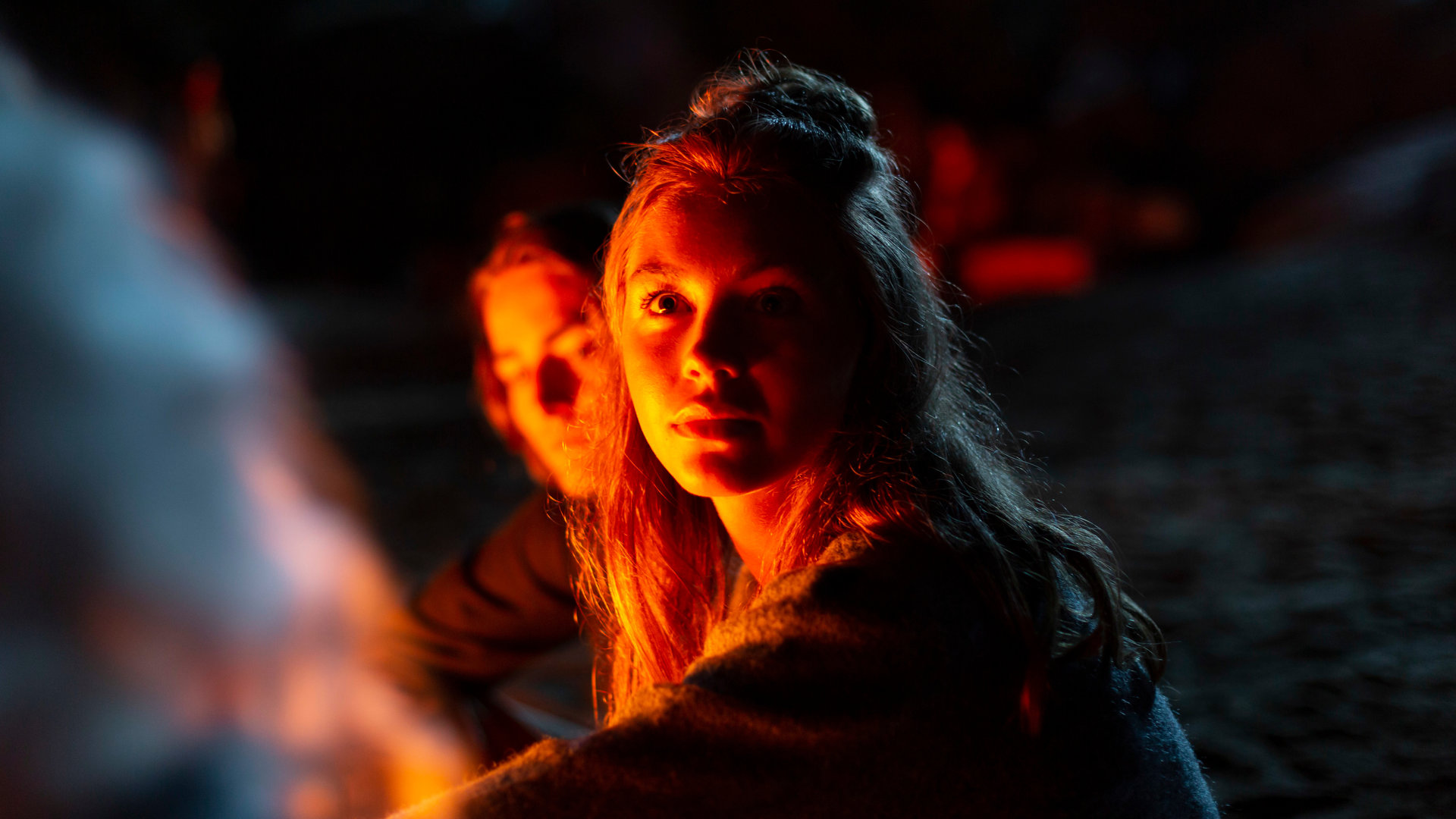 "Cryptid - Das Fest am See": Amina (Amanda Lindh) sitzt nachts mit Freunden am See. Neben ihr sitzt Ester (Maja Johanna Englander). Beide schauen sich an. Im Hintergrund sitzt Aminas Bruder Emil (Jacques Karlberg).