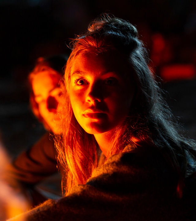 "Cryptid - Das Fest am See": Amina (Amanda Lindh) sitzt nachts mit Freunden am See. Neben ihr sitzt Ester (Maja Johanna Englander). Beide schauen sich an. Im Hintergrund sitzt Aminas Bruder Emil (Jacques Karlberg).