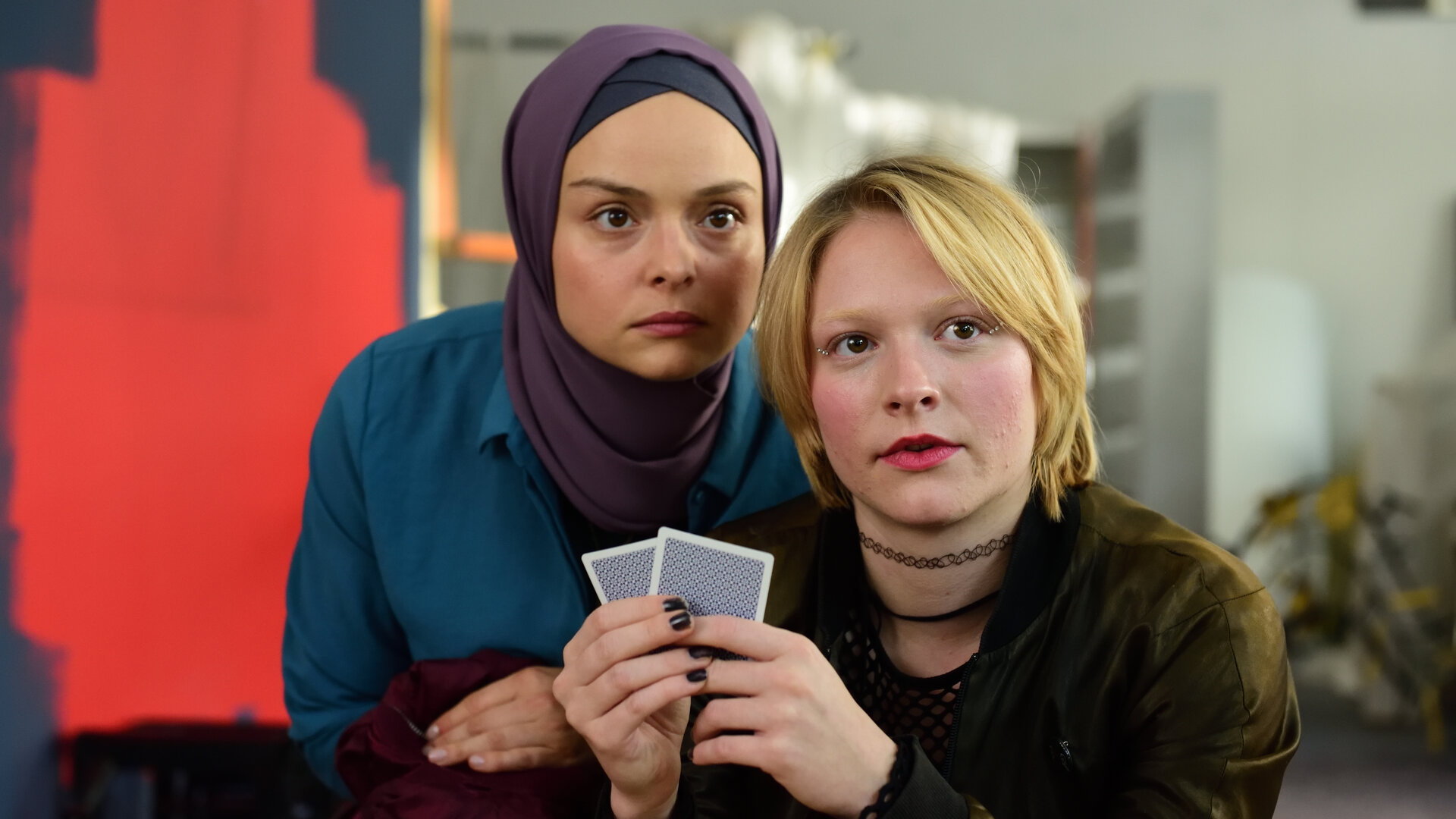 "High Stakes - Houston, I have a problem!": Britta (Pauline Großmann) hält zwei Karten vor sich und Ayla (Via Jikeli). Beide schauen nach vorne.