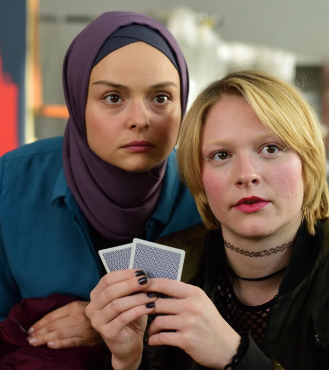 "High Stakes - Houston, I have a problem!": Britta (Pauline Großmann) hält zwei Karten vor sich und Ayla (Via Jikeli). Beide schauen nach vorne.