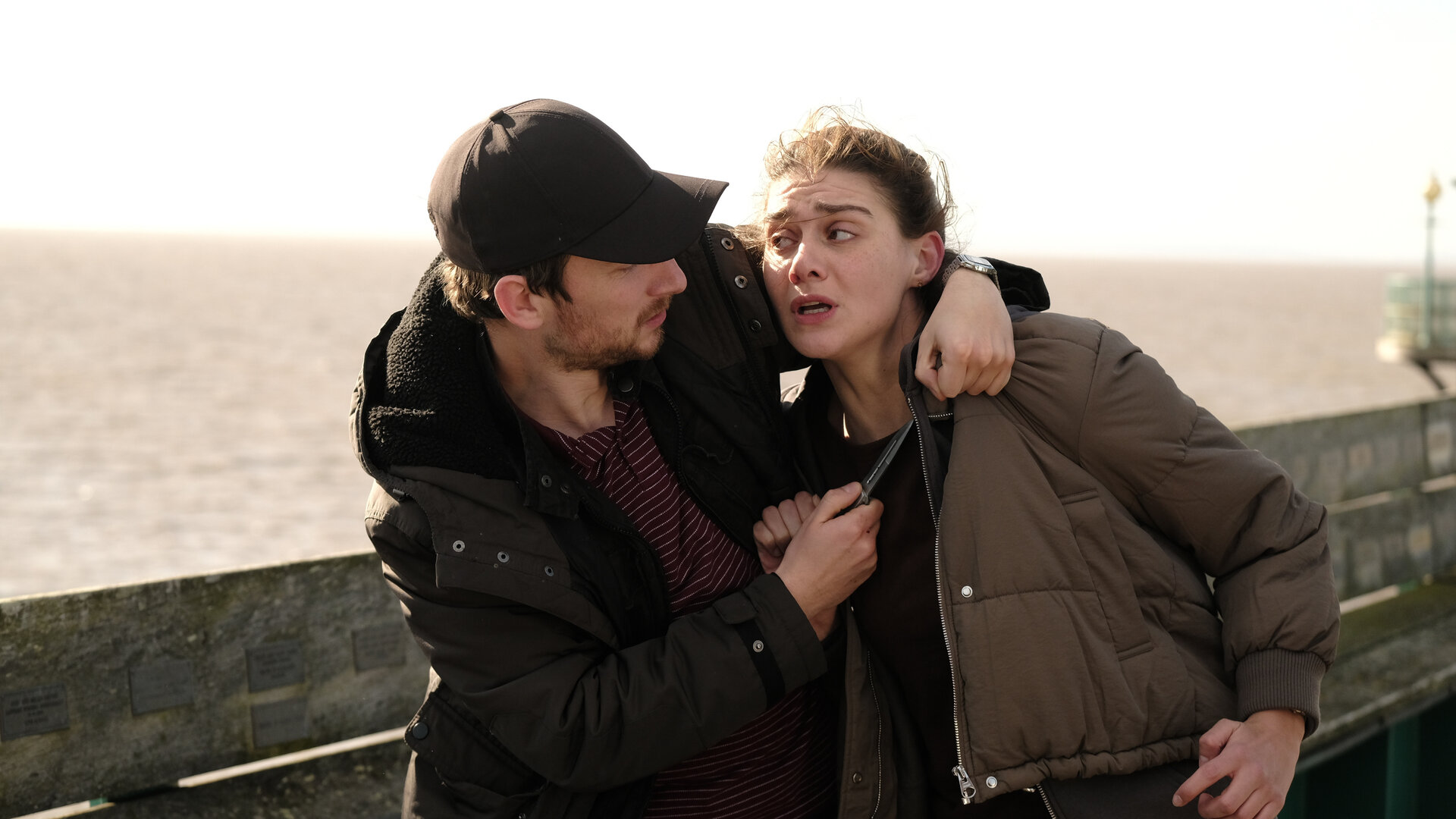 "The Killing Kind: Wolf 17": Auf einem Pier: Jake Seaton (Charles Furness) hält Ingrid (Emma Appleton) ein Messer an den Hals. Hinter ihnen verläuft eine Brüstung, dahinter liegt das Meer.