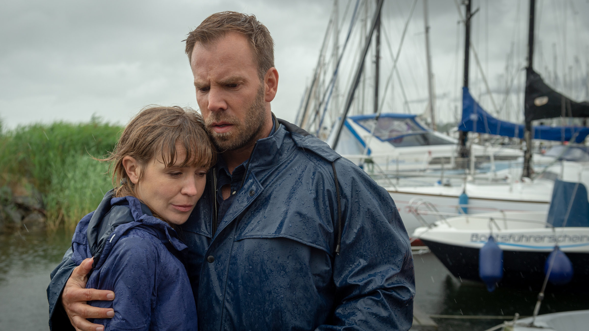 „SOKO Wismar – Tod eines Helden“: Lars Pöhlmann (Dominic Boeer) hält Astrid Lissek (Lea Faßbender) tröstend im Arm. Am Himmel hängen dunkle Wolken, es regnet in Strömen. Im Hintergrund liegen Segelboote an einem Steg.