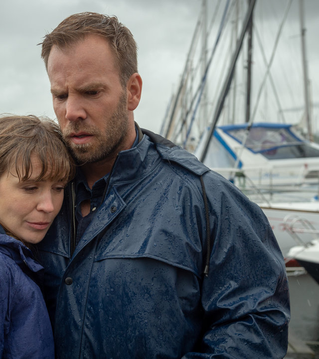 „SOKO Wismar – Tod eines Helden“: Lars Pöhlmann (Dominic Boeer) hält Astrid Lissek (Lea Faßbender) tröstend im Arm. Am Himmel hängen dunkle Wolken, es regnet in Strömen. Im Hintergrund liegen Segelboote an einem Steg.