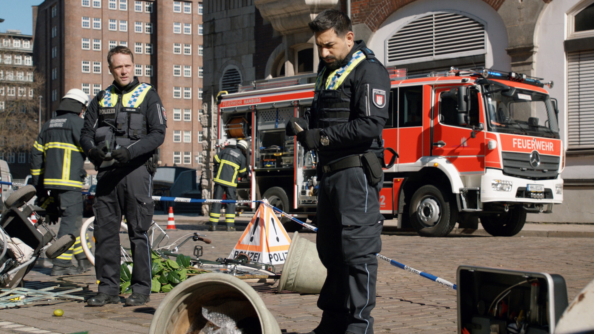 "Notruf Hafenkante - Tot geglaubt": In einer Straße mit mehrstöckigen Häusern: Die Polizisten Mattes Seeler (Matthias Schloo) und Malik Hakim (Zejhun Demirov) stehen in den Trümmern eines Unfallortes und suchen nach Hinweisen. Im Hintergrund steht ein Löschzug der Feuerwehr.