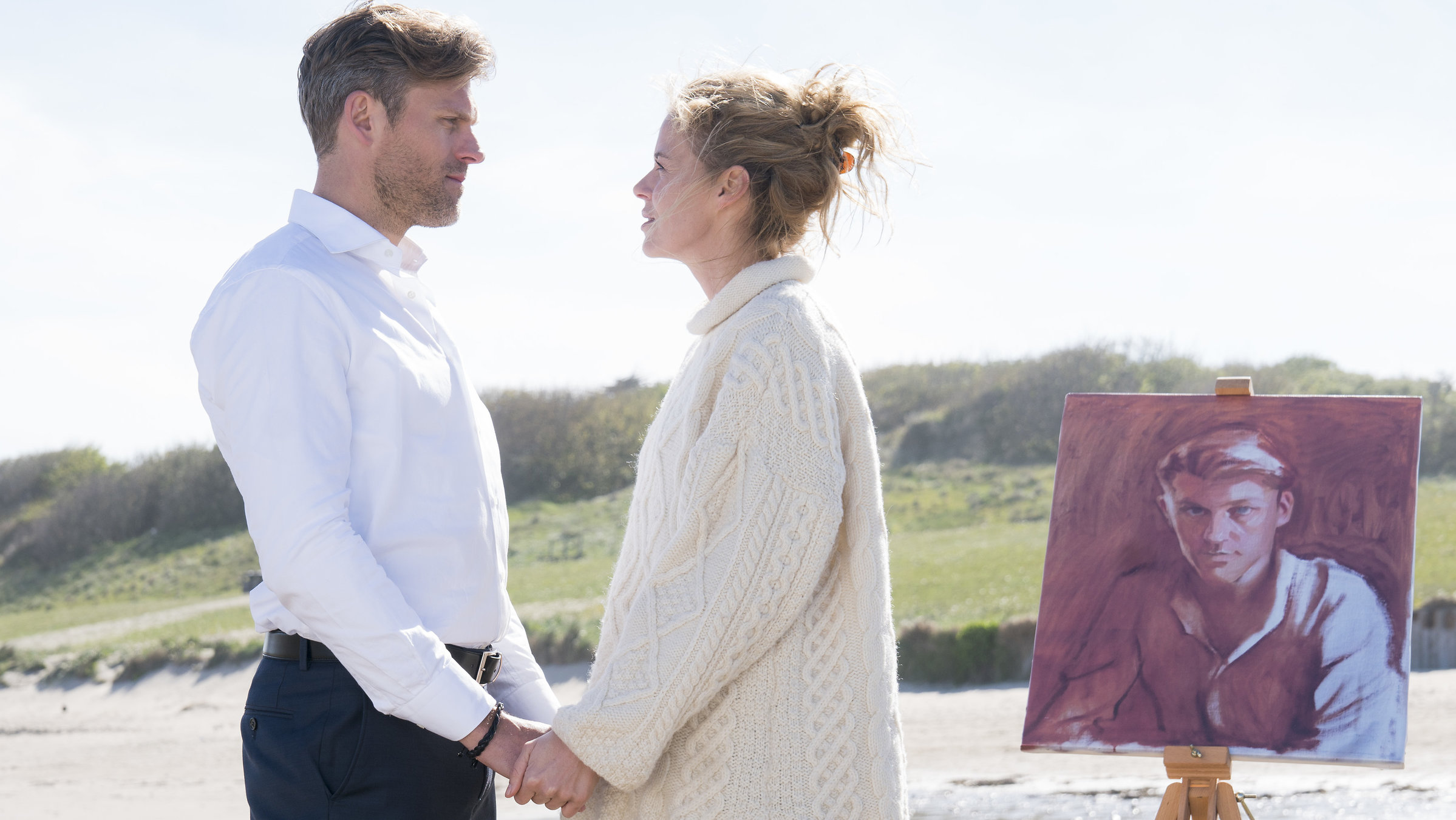 "Cecelia Ahern - Dich zu lieben": Mark (Jens Atzorn) und Claire (Jessica Ginkel) stehen Händchen haltend an einem Sandstrand und sehen sich an. Das Porträt von Mark steht auf einer Staffelei neben ihnen. Im Hintergrund sind Dünen zu erkennen.