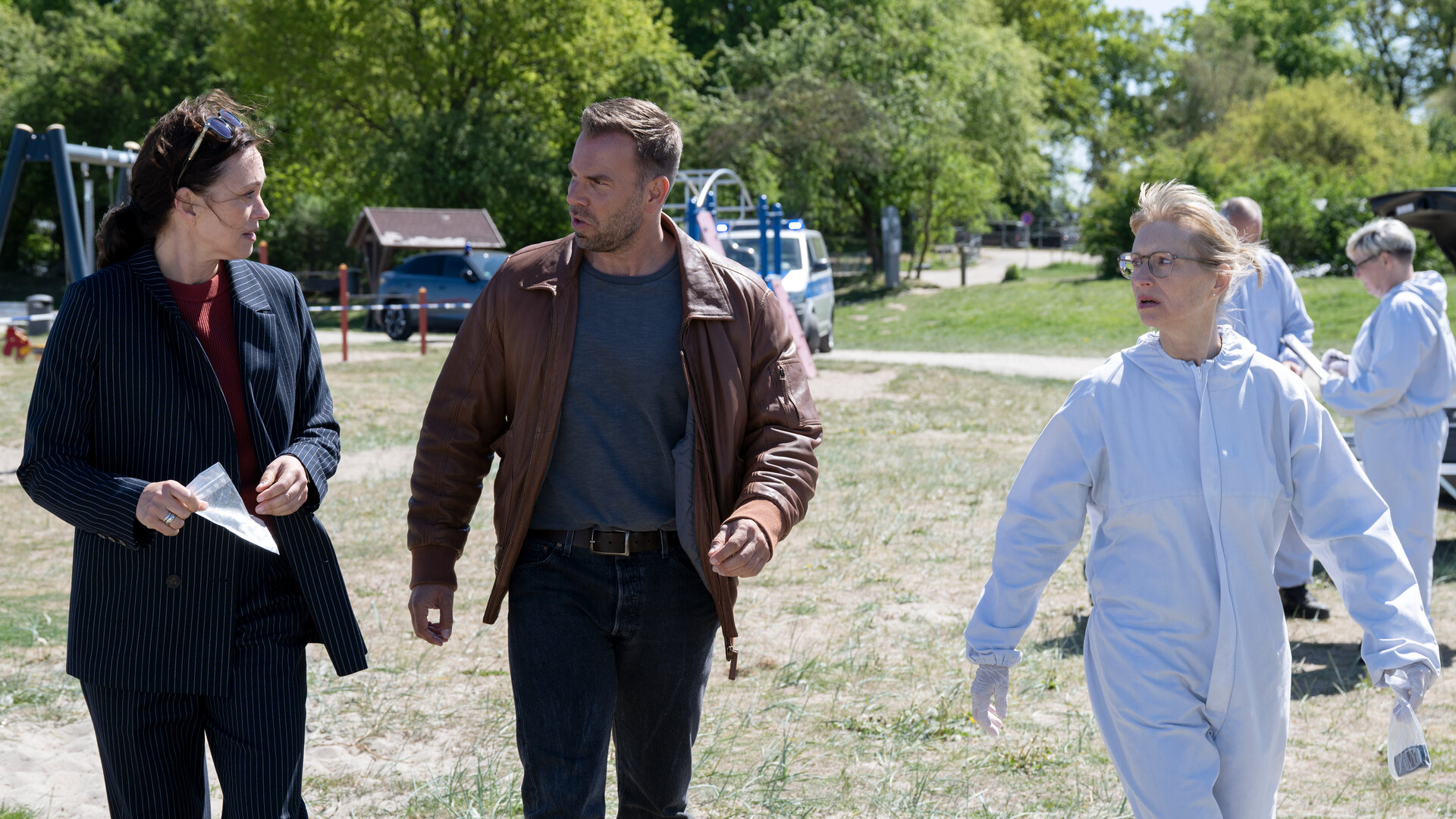 "SOKO Wismar – Der bärtige Piet": Kriminalhauptkommissarin Karoline Joost (Nike Fuhrmann) und Kriminalhauptkommissar Lars Pöhlmann (Dominic Boeer) laufen mit Kriminaltechnikerin Roswitha Prinzler (Silke Matthias) über einen Spielplatz auf den Tatort zu. Im Hintergrund arbeiten zwei Kollegen (Komparsen) der Spurensicherung.
