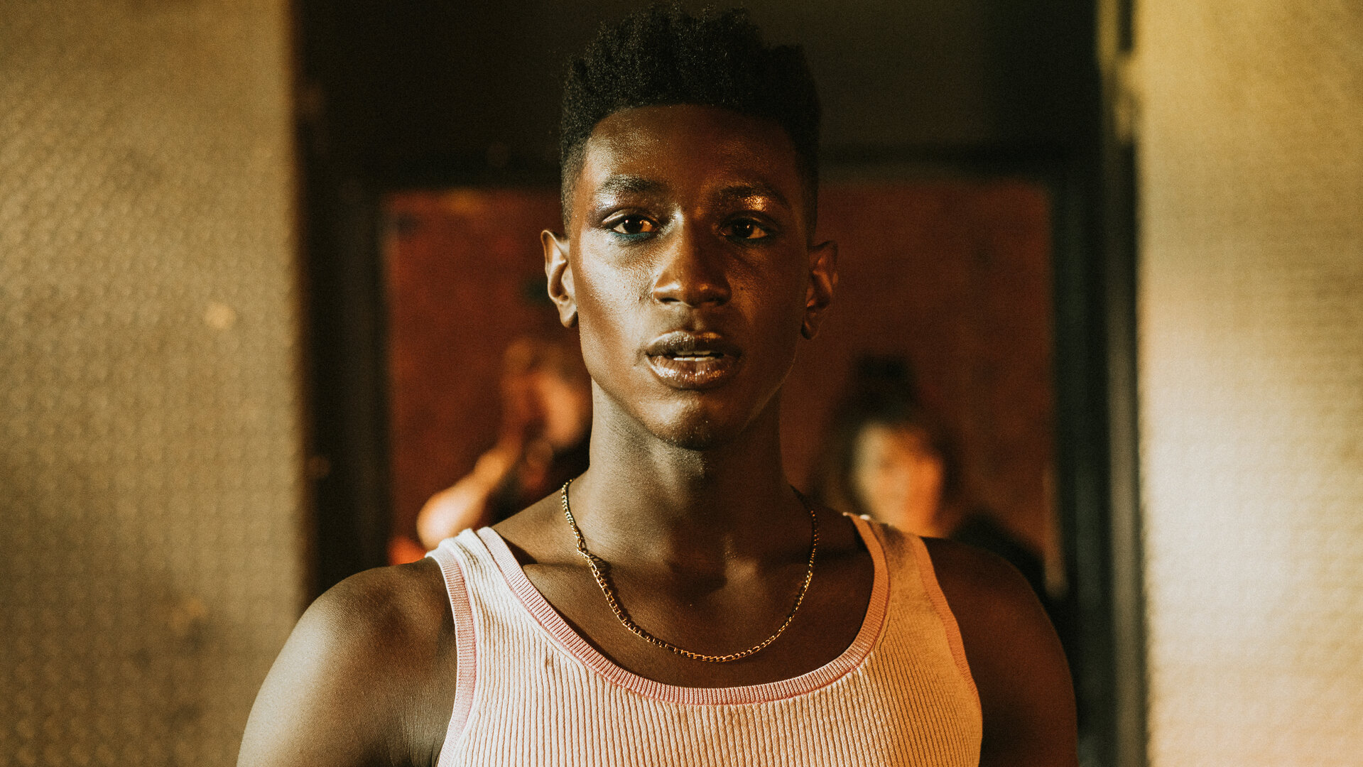 "House of Bellevue - Runway": Emm (Ricco-Jarret Boateng) steht in einem Raum vor einem dunklen Türrahmen. Er trägt ein Unterhemd und eine Goldkette.