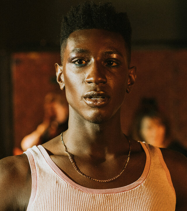 "House of Bellevue - Runway": Emm (Ricco-Jarret Boateng) steht in einem Raum vor einem dunklen Türrahmen. Er trägt ein Unterhemd und eine Goldkette.