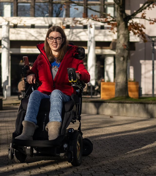 "einfach Mensch - Johanna Ruhrbruch: Die rollende Reporterin": Johanna Ruhrbruch vor einer Universität in einer roten Winterjacke.