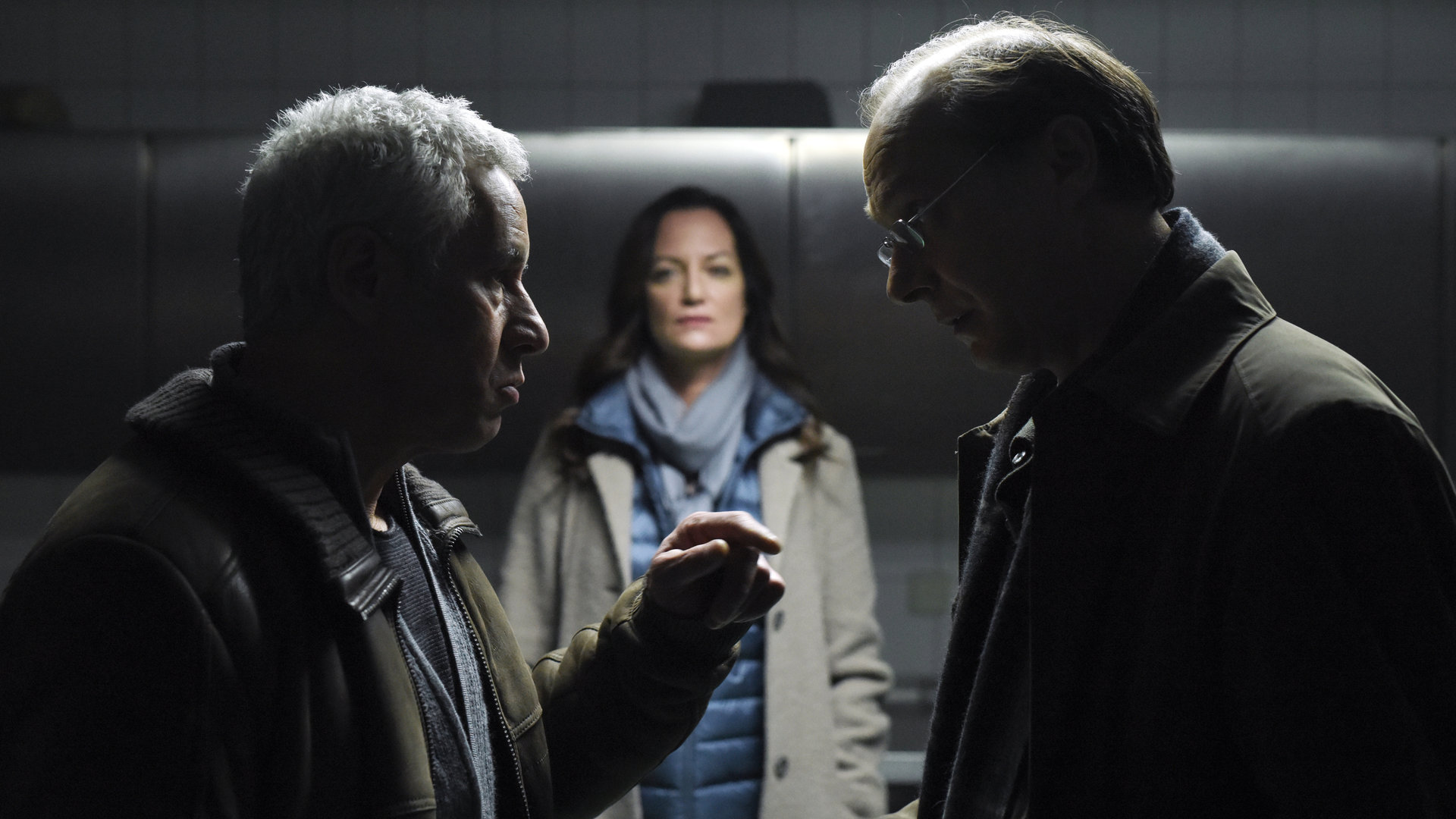 "Unter anderen Umständen – Das Geheimnis der Schwestern": Matthias Hamm (Ralph Herforth) und Arne Brauner (Martin Brambach) stehen sich in einem Raum gegenüber und unterhalten sich. Etwas im Hintergrund steht Jana Winter (Natalia Wörner) und beobachtet die beiden.