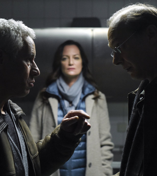 "Unter anderen Umständen – Das Geheimnis der Schwestern": Matthias Hamm (Ralph Herforth) und Arne Brauner (Martin Brambach) stehen sich in einem Raum gegenüber und unterhalten sich. Etwas im Hintergrund steht Jana Winter (Natalia Wörner) und beobachtet die beiden.