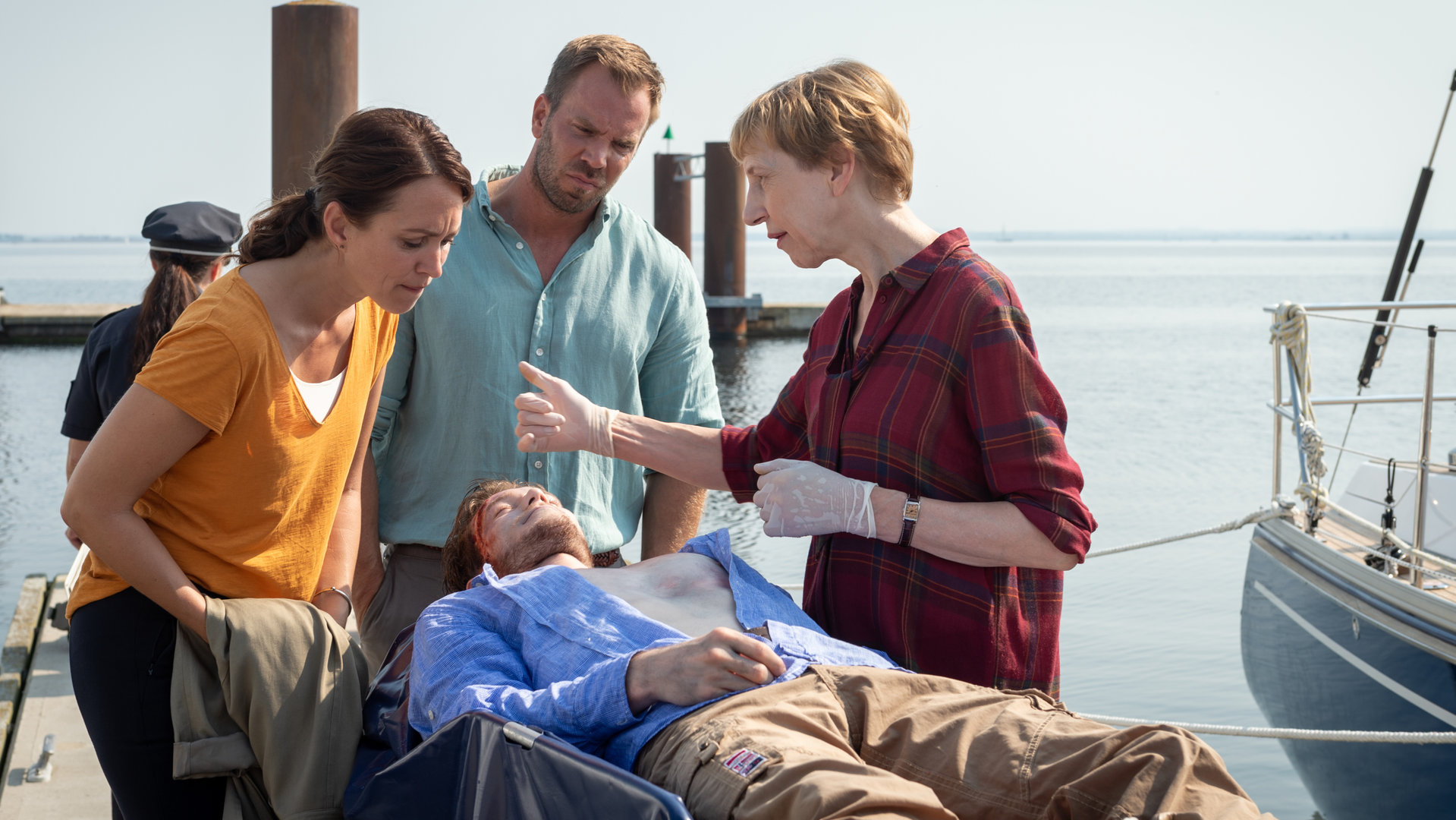 „SOKO Wismar – Wann, wenn nicht jetzt?“: Ole Janisch (Max Brauer) liegt tot auf einer Trage. Um ihn herum stehen Helene Sturbeck (Katharina Blaschke), Lars Pöhlmann (Dominic Boeer) und Karoline Joost (Nike Fuhrmann). Helene deutet auf die blutige Wunde an der Stirn des Toten. Im Hintergrund liegt das offene Meer.