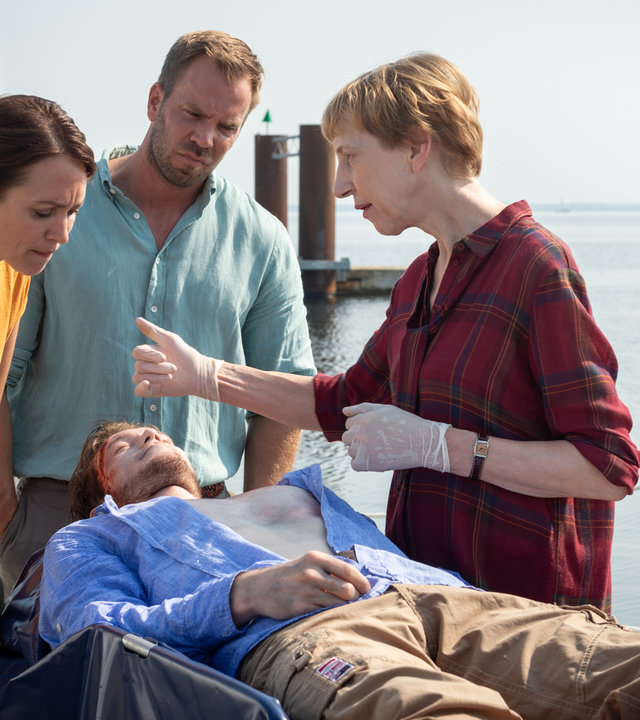 „SOKO Wismar – Wann, wenn nicht jetzt?“: Ole Janisch (Max Brauer) liegt tot auf einer Trage. Um ihn herum stehen Helene Sturbeck (Katharina Blaschke), Lars Pöhlmann (Dominic Boeer) und Karoline Joost (Nike Fuhrmann). Helene deutet auf die blutige Wunde an der Stirn des Toten. Im Hintergrund liegt das offene Meer.