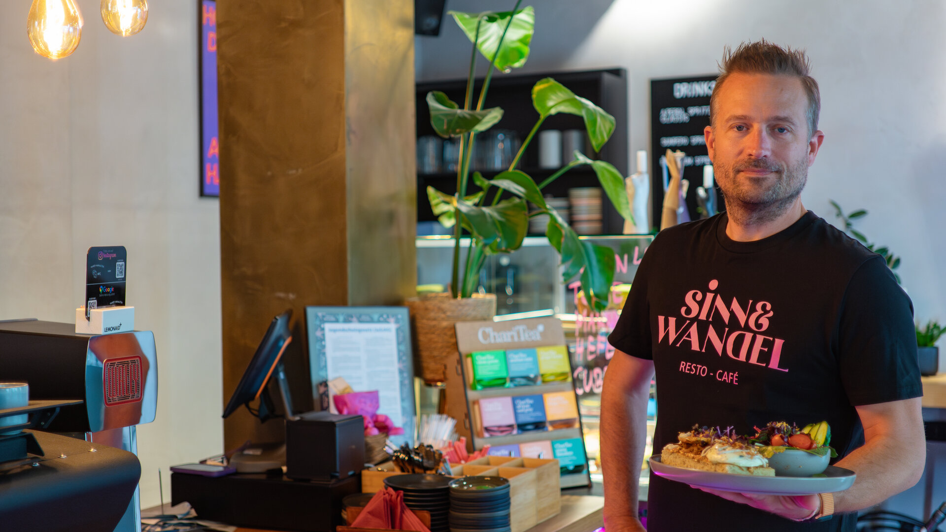 "einfach Mensch - Sascha Nuhn: Ein Gastronom findet Gehör": Der Protagonist Sascha Nuhn steht mit einem Teller in der Hand vor der Theke in seinem Café "Sinn&Wandel".