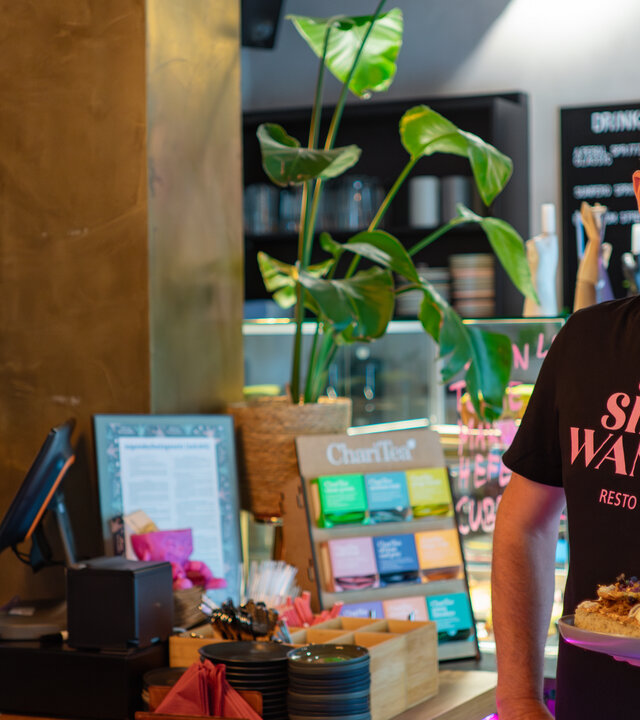 "einfach Mensch - Sascha Nuhn: Ein Gastronom findet Gehör": Der Protagonist Sascha Nuhn steht mit einem Teller in der Hand vor der Theke in seinem Café "Sinn&Wandel".