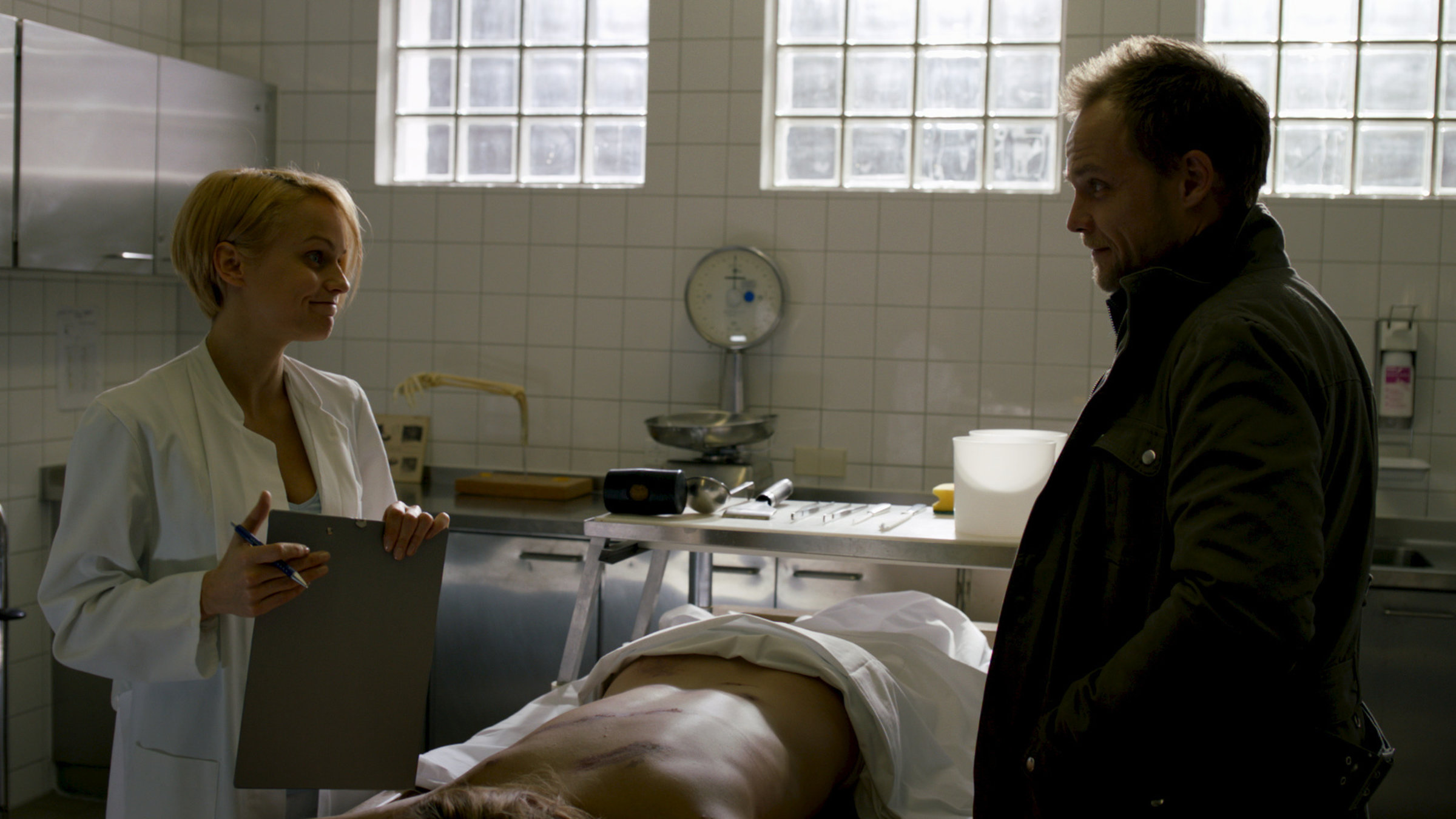 "Kommissar Marthaler - Die Braut im Schnee": In der Gerichtsmedizin steht Dr. Thea Hollmann (Mirjam Weichselbraun) neben Kommissar Robert Marthaler (Matthias Koeberlin). Zwischen den beiden liegt eine Leiche auf einer Bare.