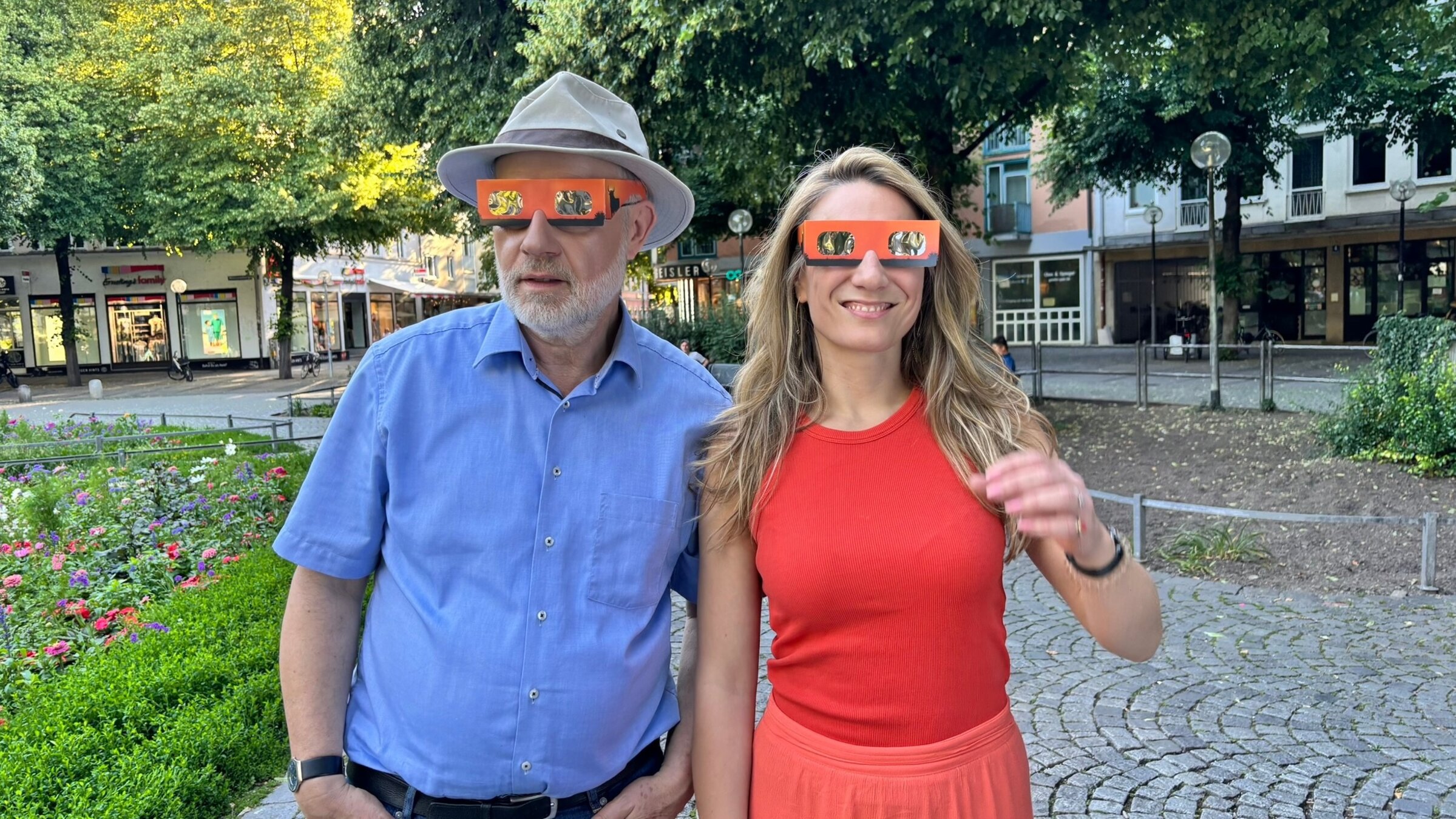 "Sonnenfinsternis über Mallorca": Harald Lesch und Jasmina Neudecker tragen beide Sonnenfinsternis-Brillen. Sie stehen in einem kleinen städtischen Park.